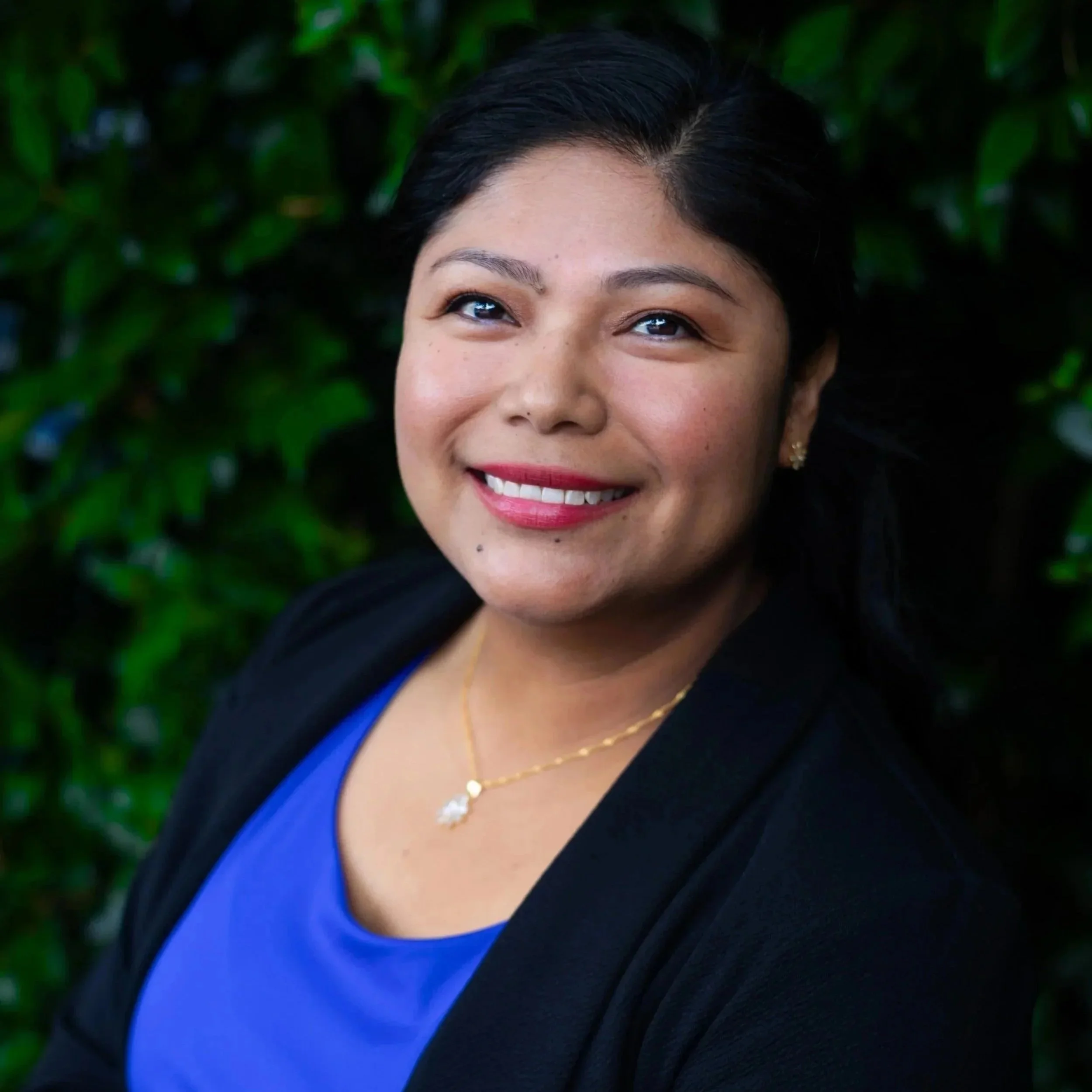 A portrait of a woman with dark hair, smiling, wearing a blue top and a black blazer, with a gold necklace, posing outdoors with green foliage in the background.
