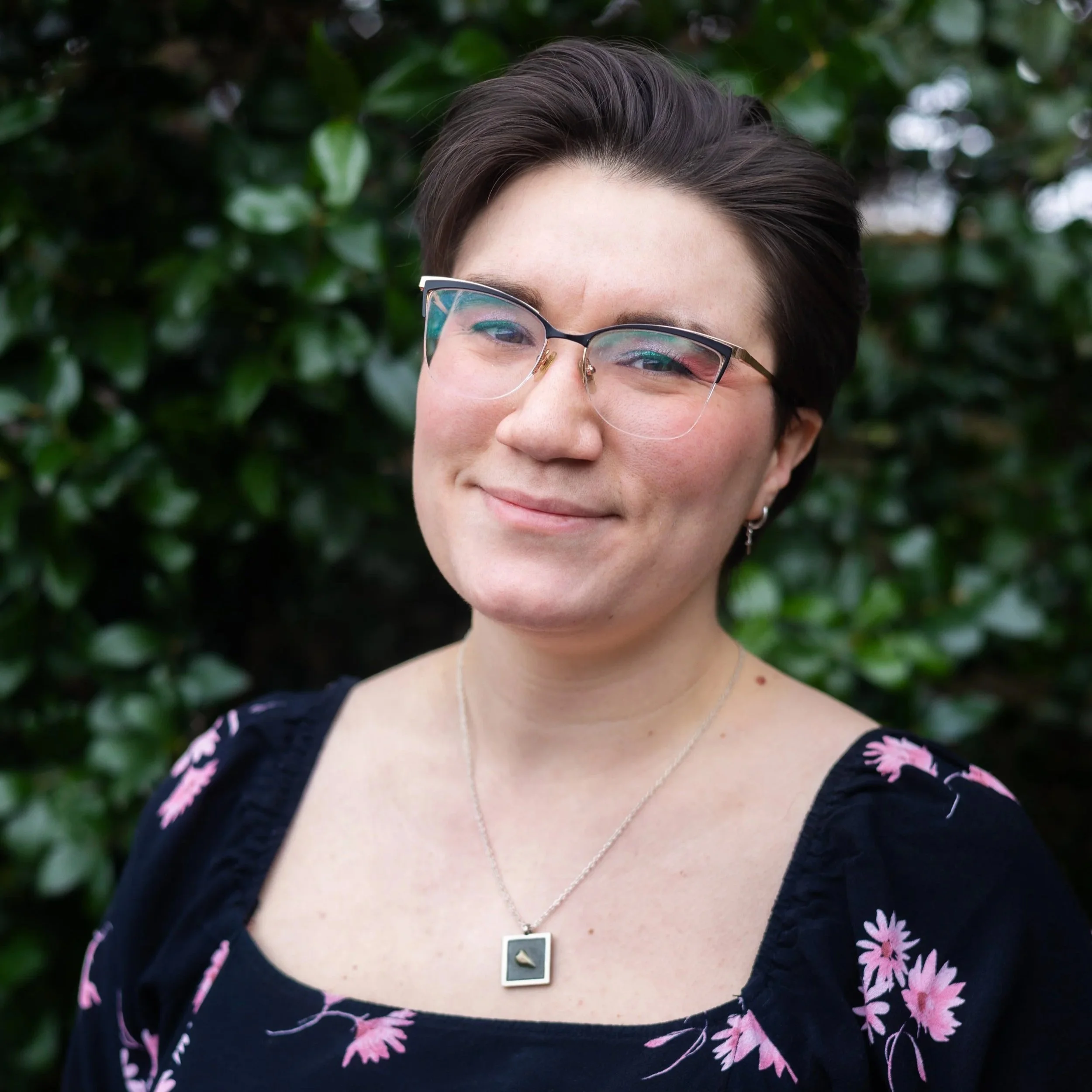 A young woman with short dark hair, wearing glasses with a thin frame, a black dress with pink floral patterns, and a silver necklace with a small pendant, smiling in front of a background of green leaves.