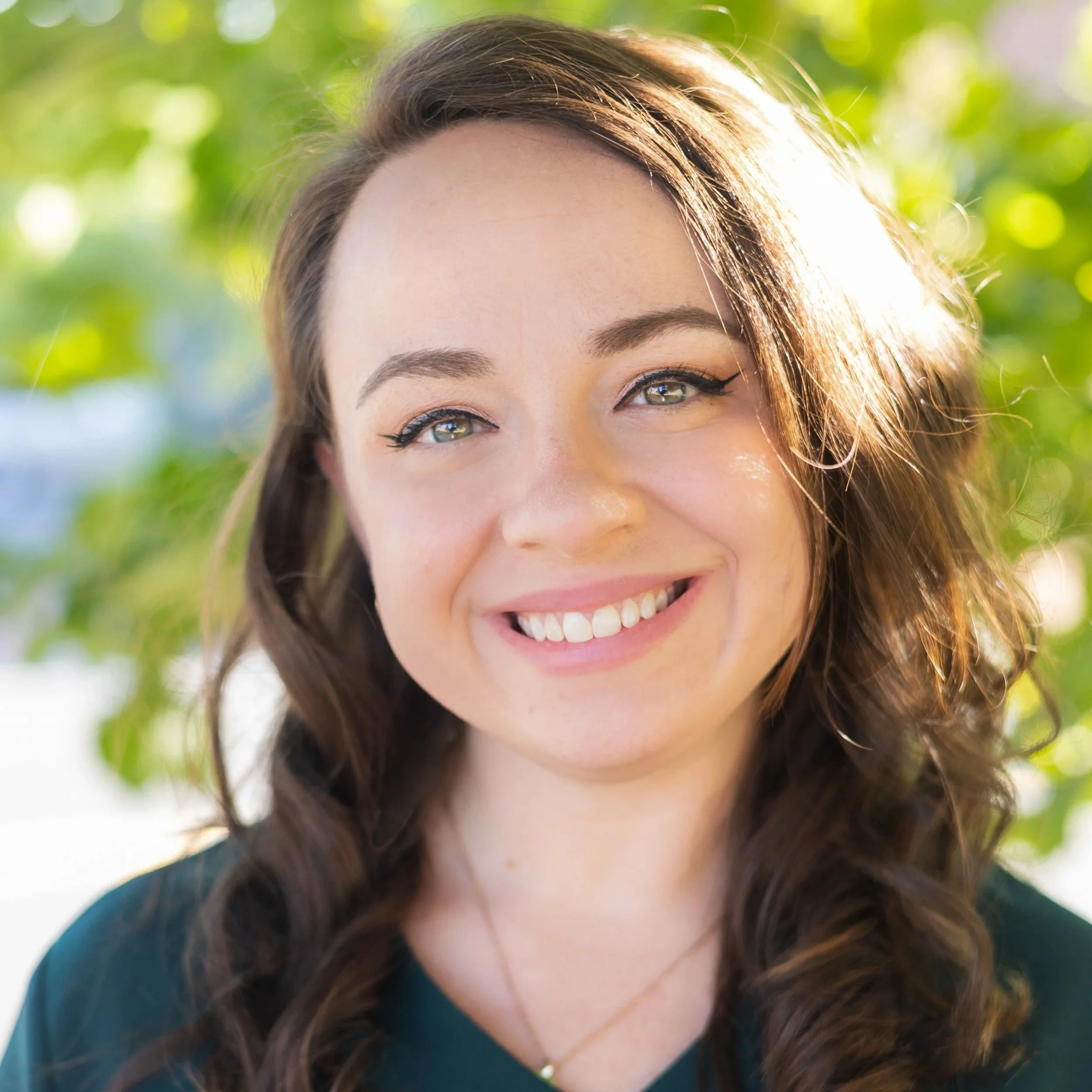 Close-up portrait of a smiling young woman with wavy brown hair, green eyes, and makeup, outdoors with blurred green foliage in the background.