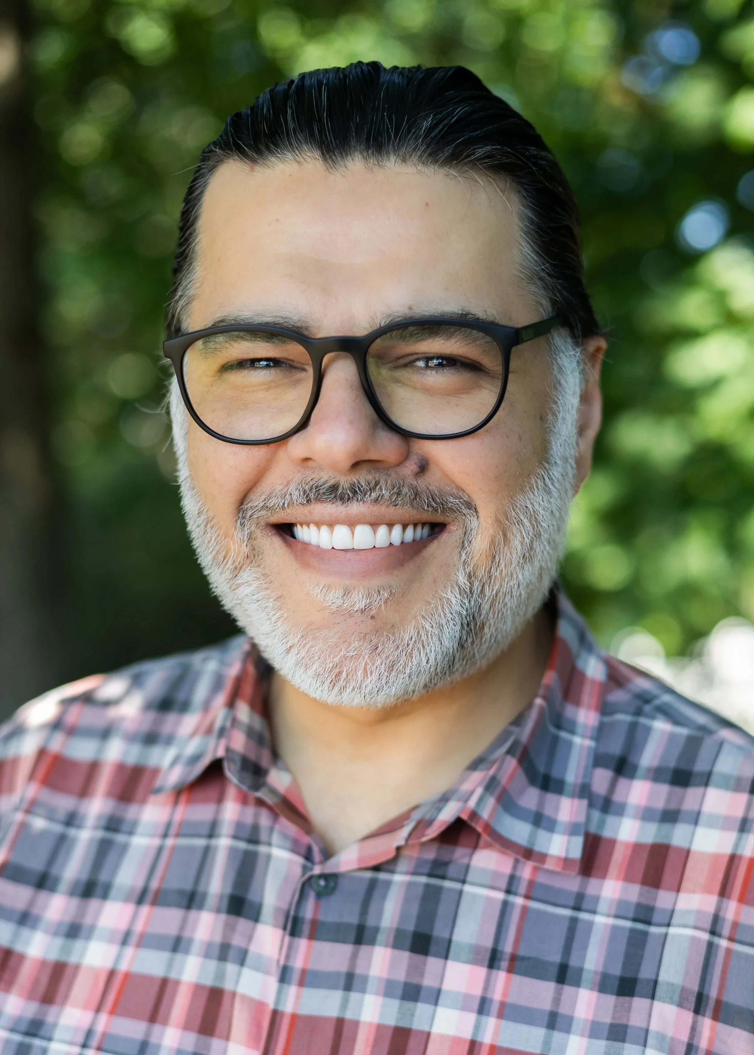 Close-up of a smiling middle-aged man with glasses, salt-and-pepper hair and beard, wearing a plaid shirt, outdoors with green trees in the background.
