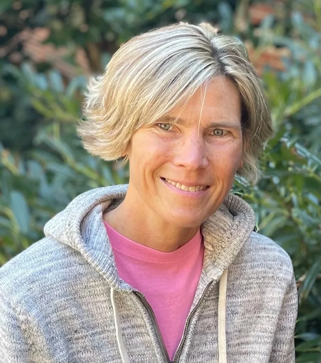 A woman with short blonde hair and blue eyes smiling, wearing a pink shirt and a light gray zip-up hoodie, standing outdoors with green foliage in the background.