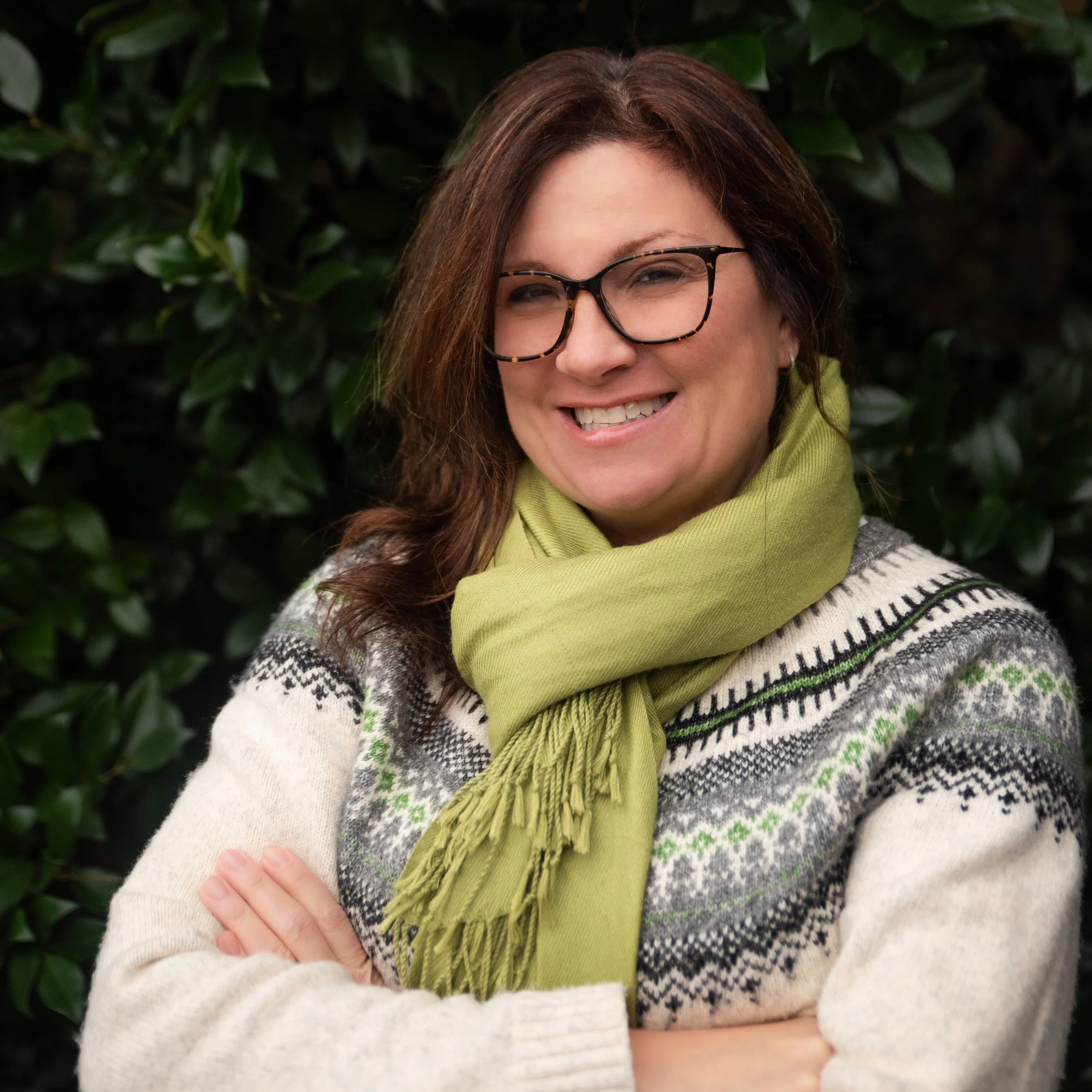 A woman with brown hair, glasses, and a green scarf, smiling with arms crossed in front of a leafy background.