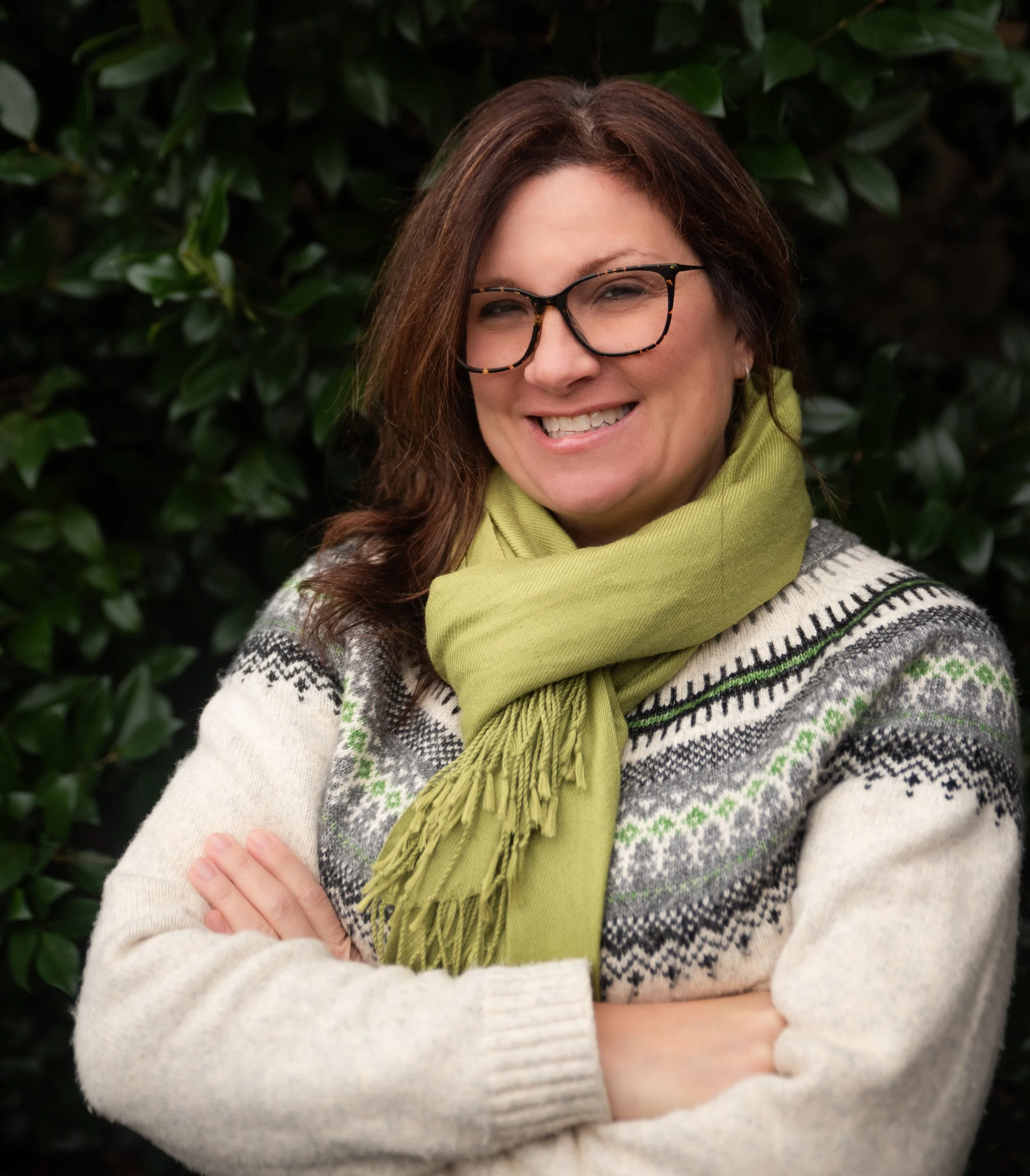 Woman with dark brown hair, glasses, smiling, wearing a cream-colored sweater with a Nordic pattern and a green scarf, standing outdoors in front of green foliage.