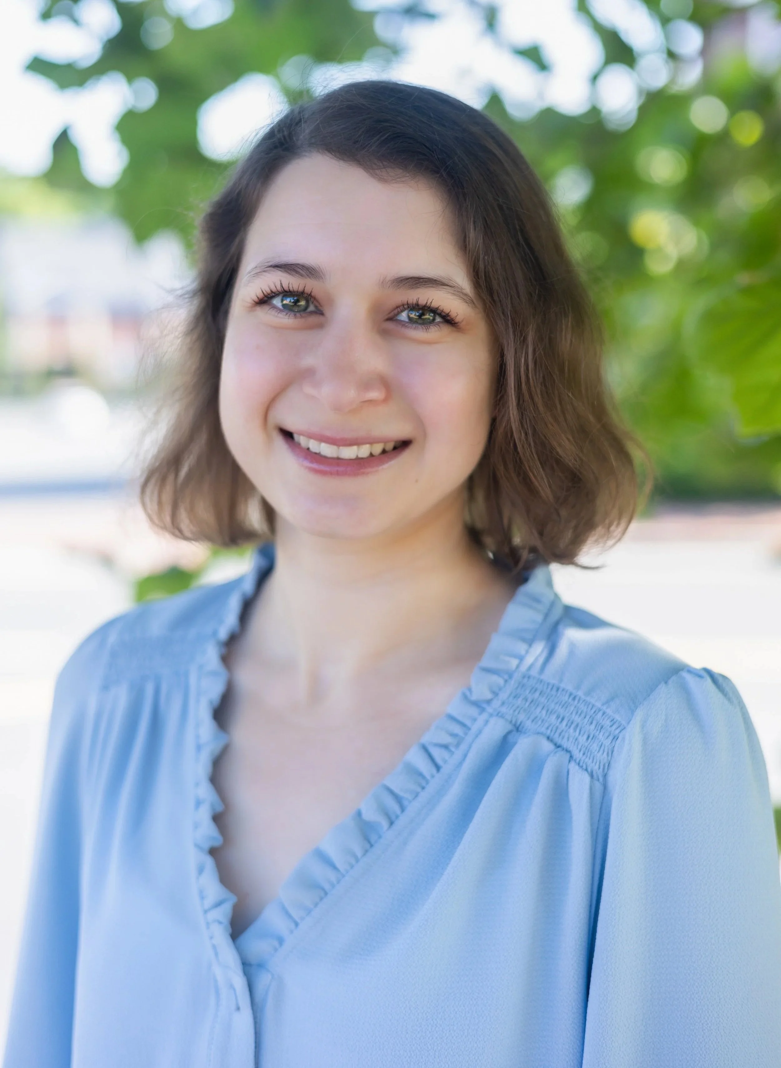 A young woman with short brown hair and blue eyes smiling outdoors, wearing a light blue blouse with ruffled collar, with green trees in the background.