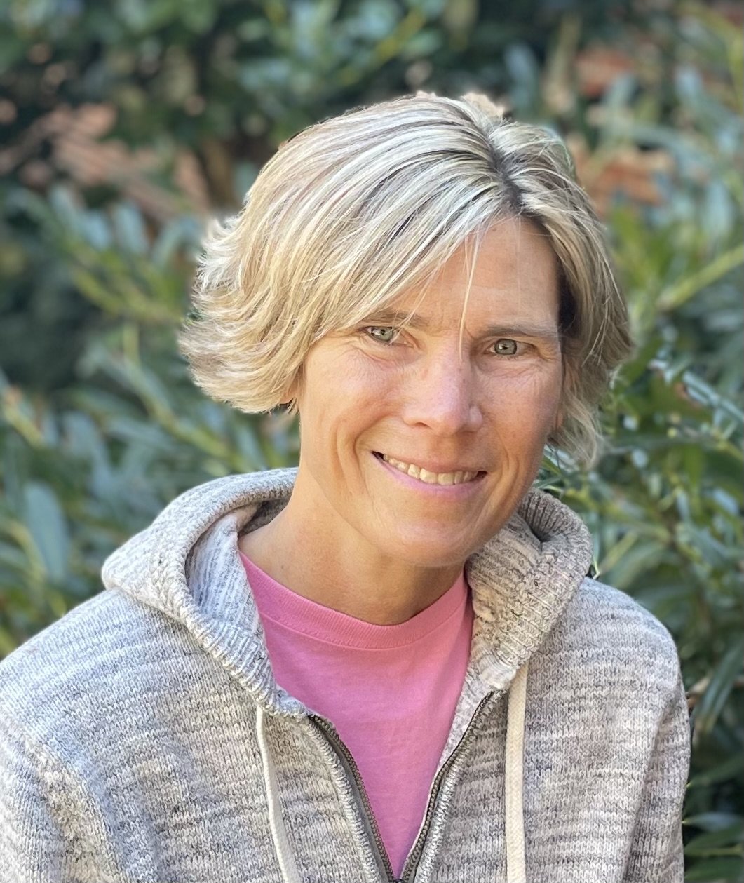 A woman with short blonde hair wearing a pink shirt and gray hoodie, standing outdoors with green foliage in the background.