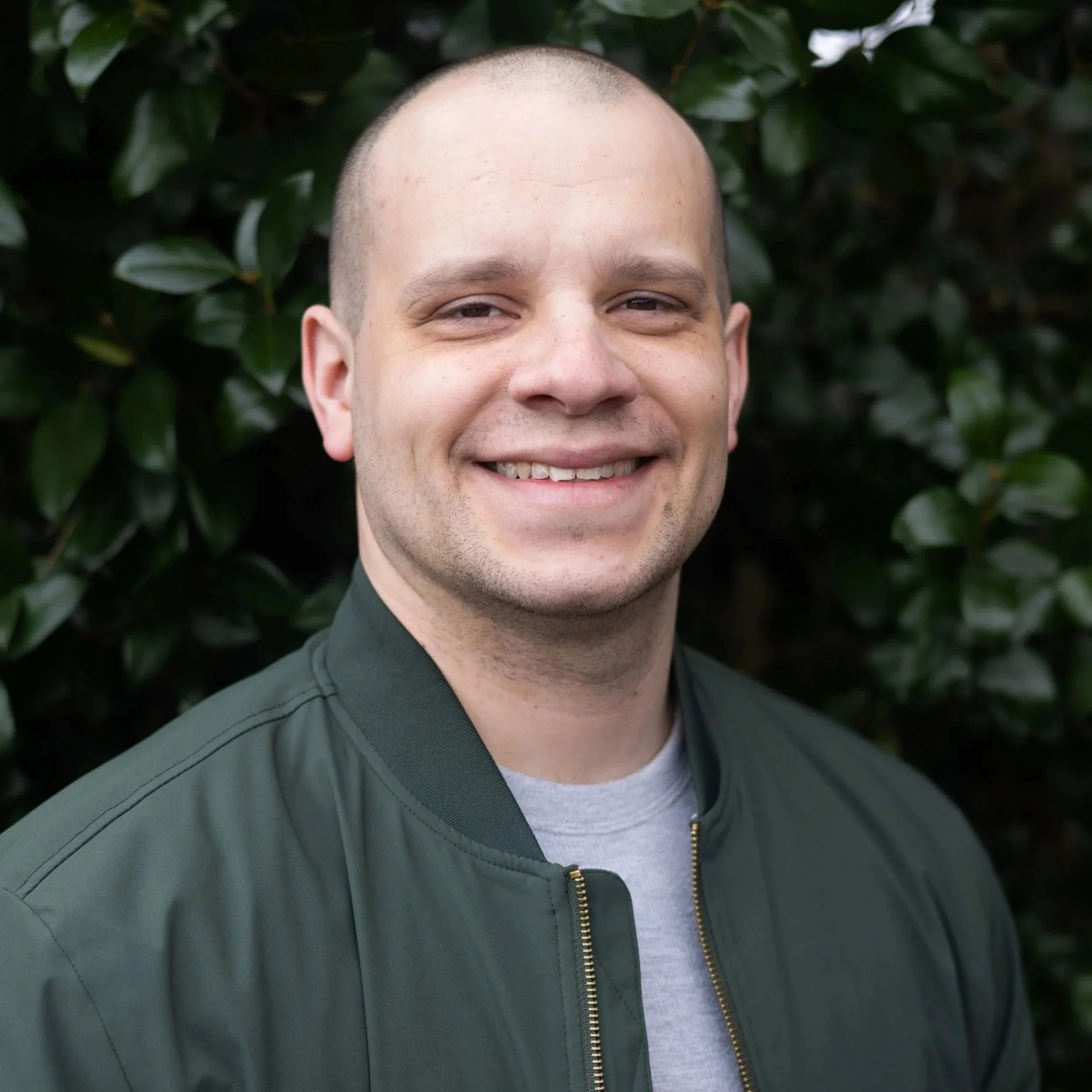 A smiling man with a shaved head wearing a grey t-shirt and a green military-style jacket, standing outdoors in front of a leafy green bush.
