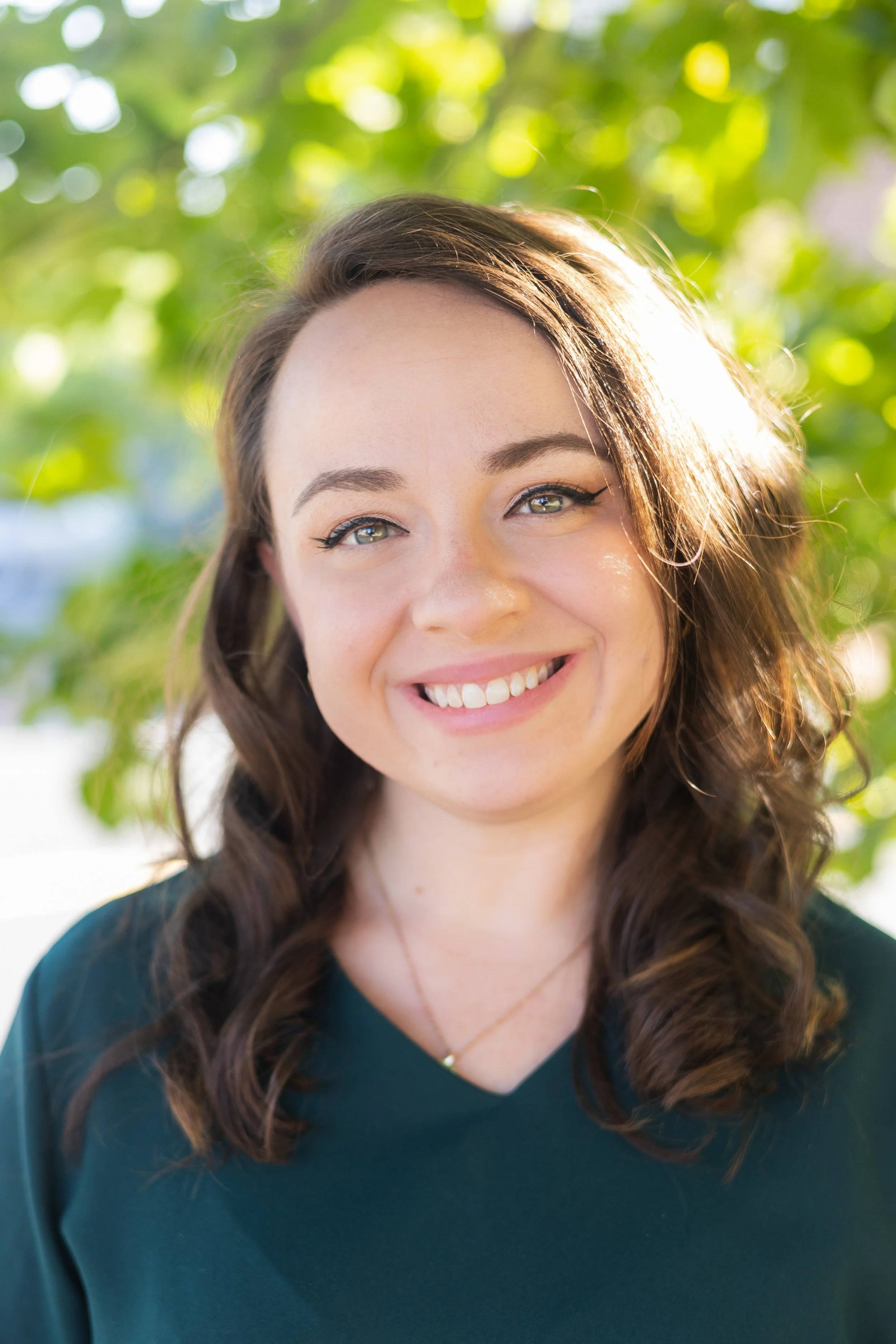 A young woman with wavy brown hair, blue eyes, and light skin, smiling outdoors with sunlight filtering through green leaves in the background.
