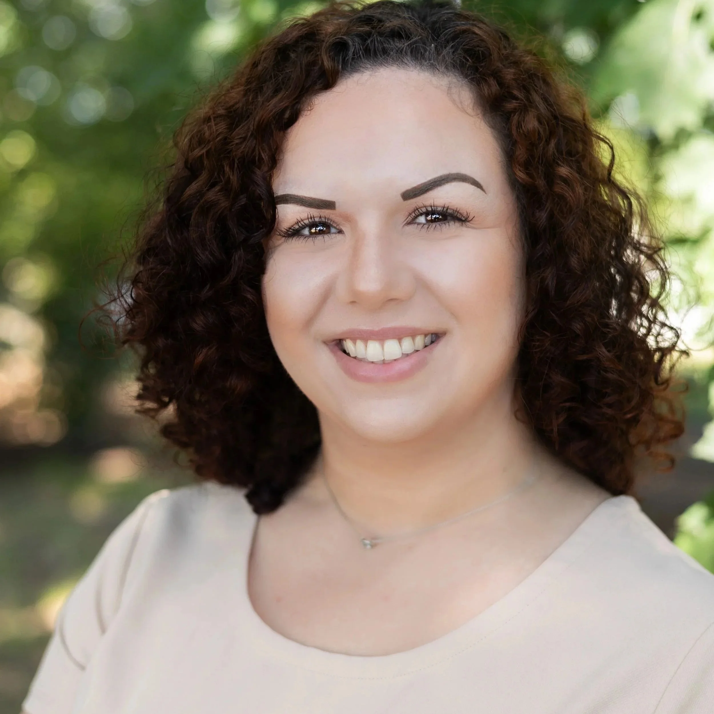 Close-up of a smiling woman with curly brown hair, wearing a beige top and a delicate necklace, outdoors with blurred green trees in the background.