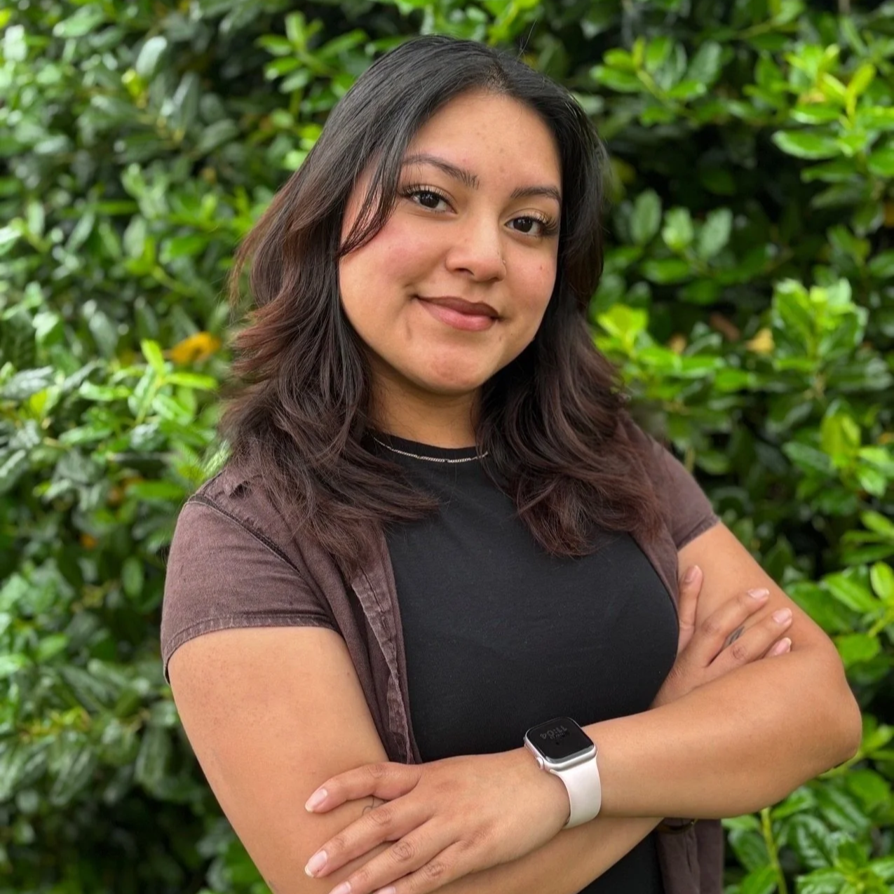 A young woman with brown, wavy hair standing in front of green foliage, smiling with arms crossed, wearing a black shirt, brown jacket, and a white smartwatch.