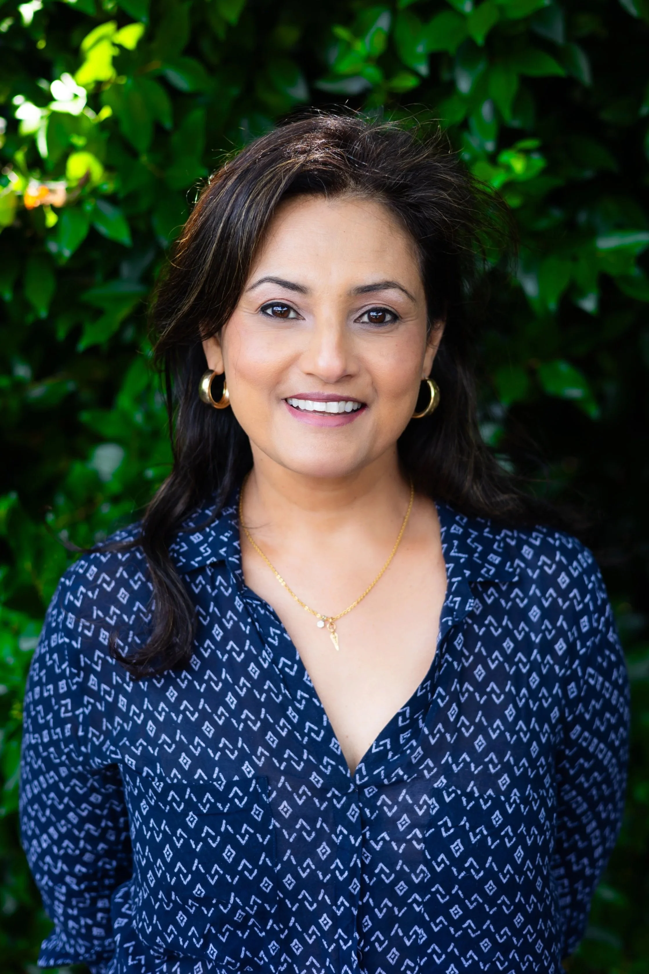 A woman with dark hair and gold jewelry, smiling, outdoors with green leafy background.