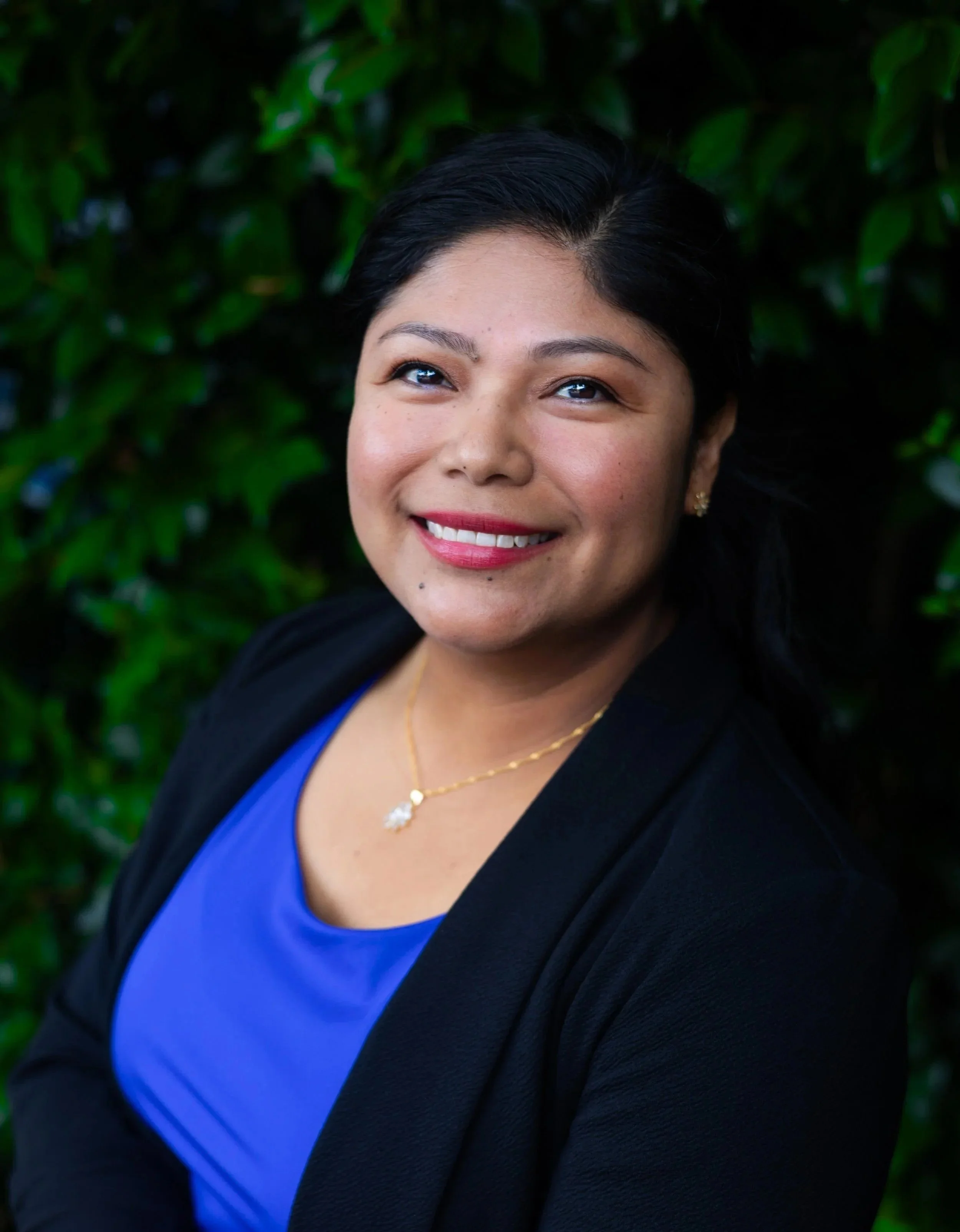 A woman smiling, wearing a blue top, black blazer, and gold jewelry, standing in front of green foliage.