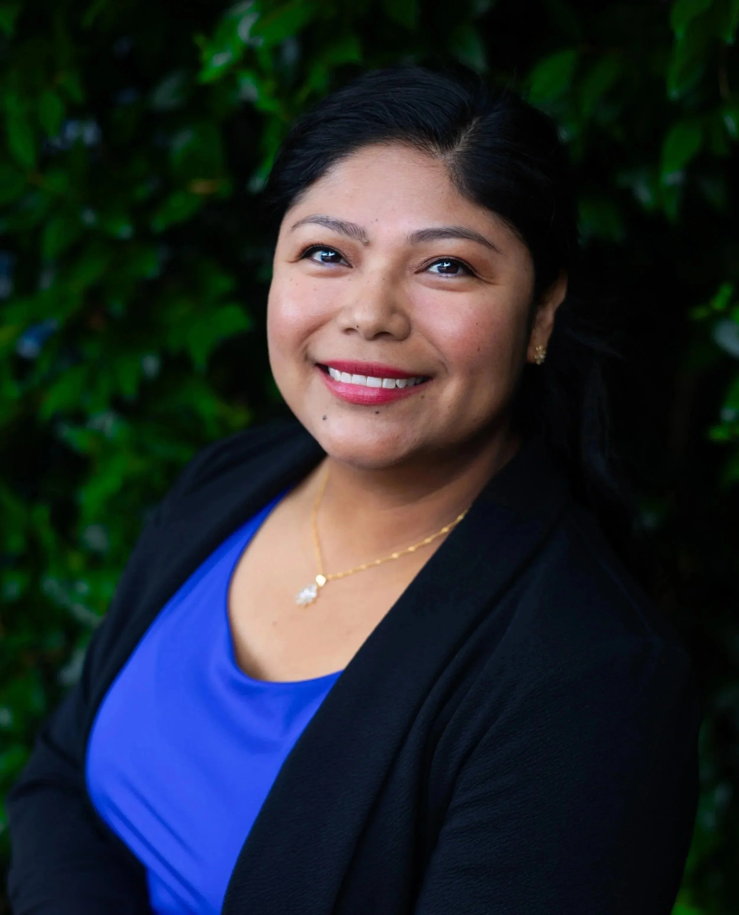 A woman with black hair wearing a black blazer and a blue top, smiling, with a background of green leaves.