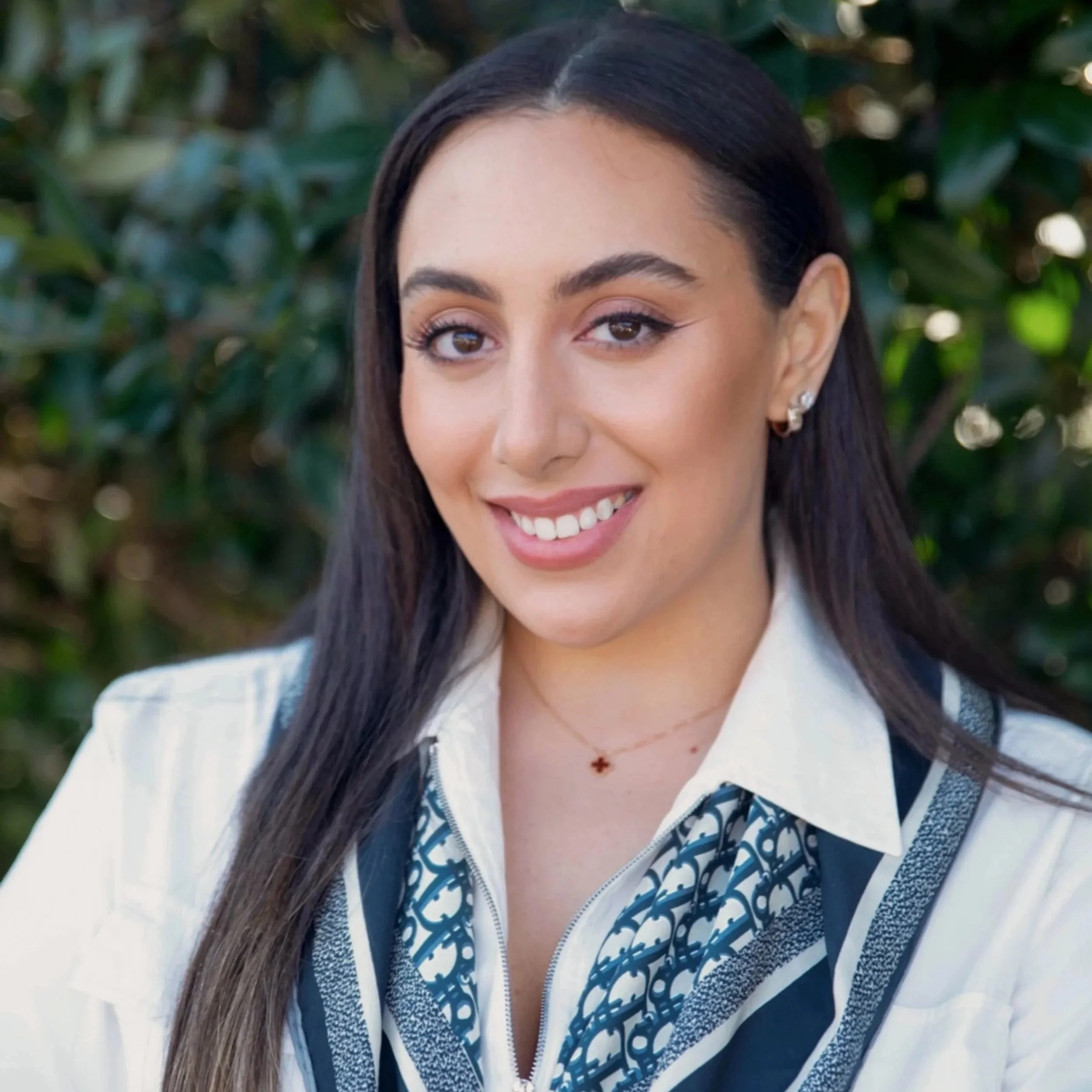 Portrait of a woman with long dark hair, smiling, wearing earrings, a necklace with a small pendant, a white shirt, and a patterned scarf, outdoors with greenery in the background.