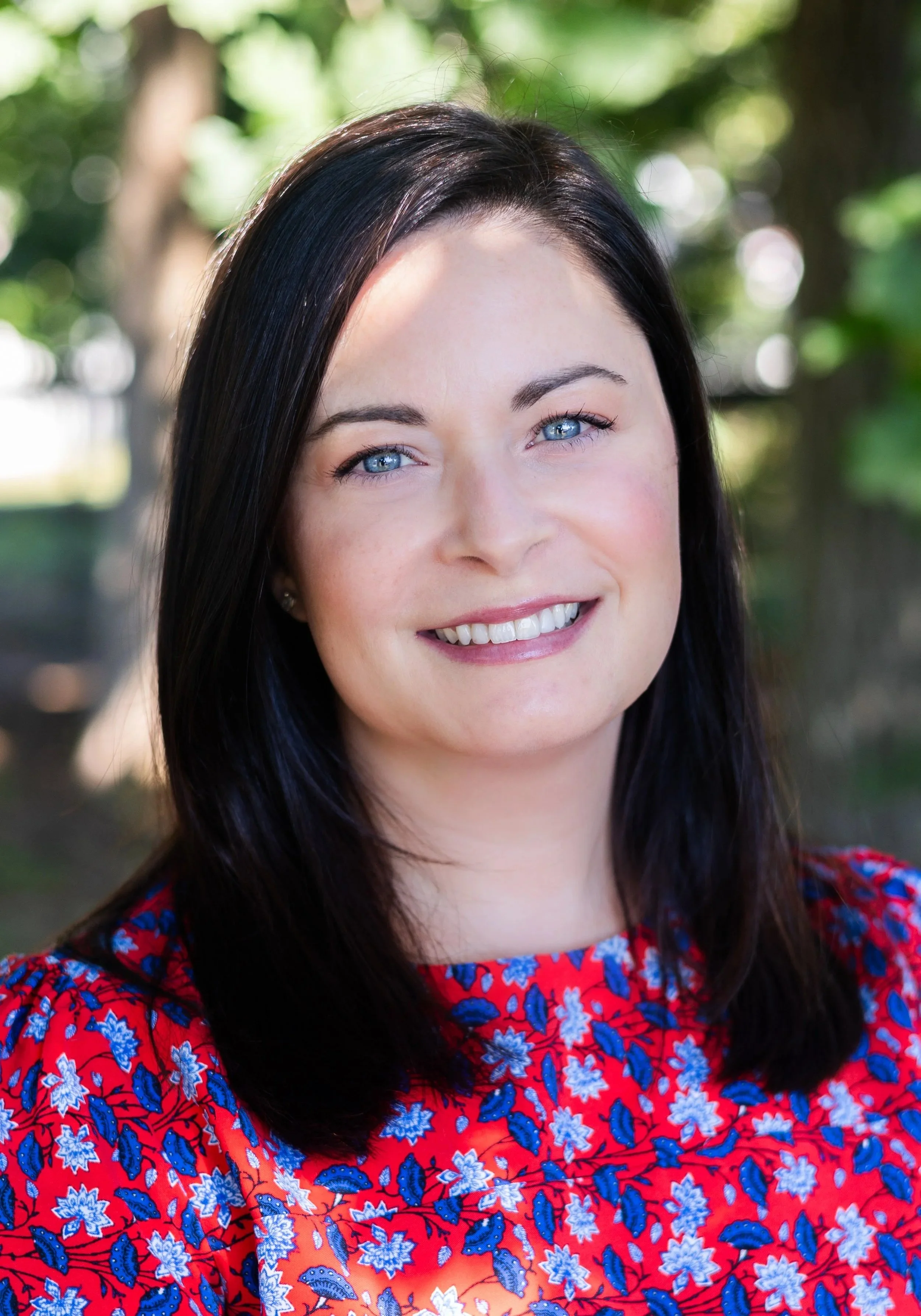A woman with blue eyes and black hair smiling outdoors, wearing a red floral dress.