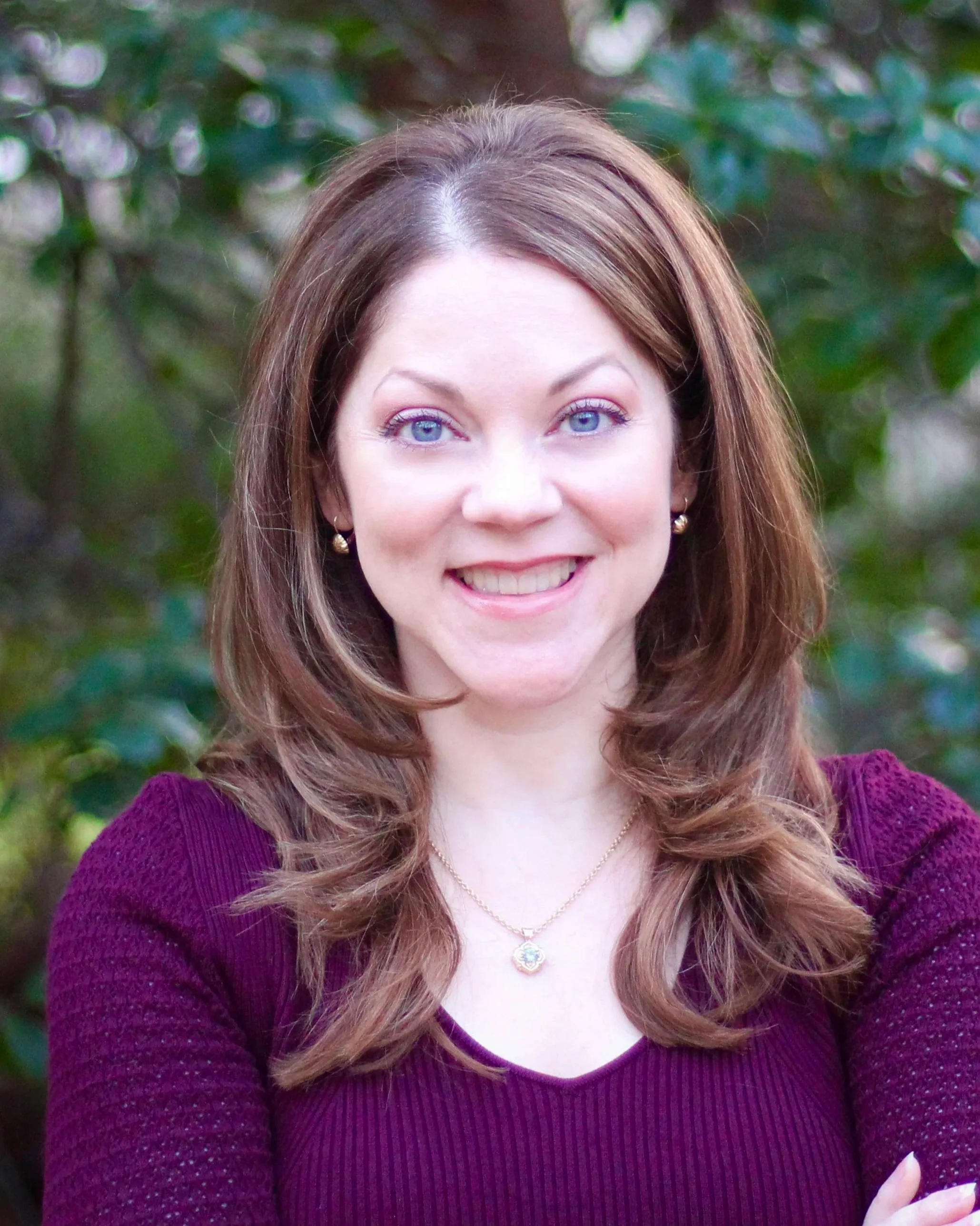 A woman with blue eyes, smiling, outdoors with green foliage in the background, wearing a maroon sweater and gold jewelry.