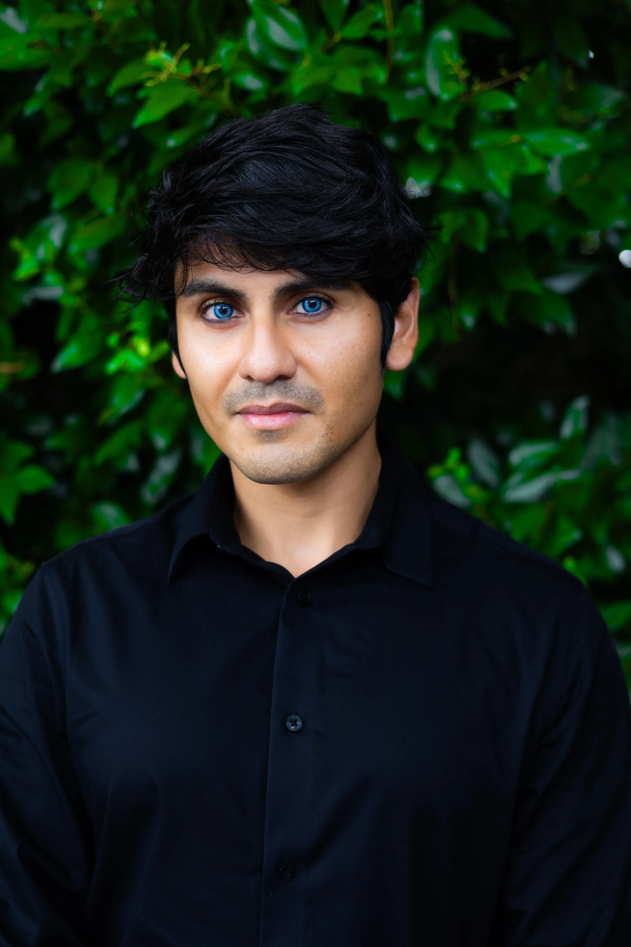 A man with bright blue eyes and dark hair wearing a black shirt, standing in front of green foliage.