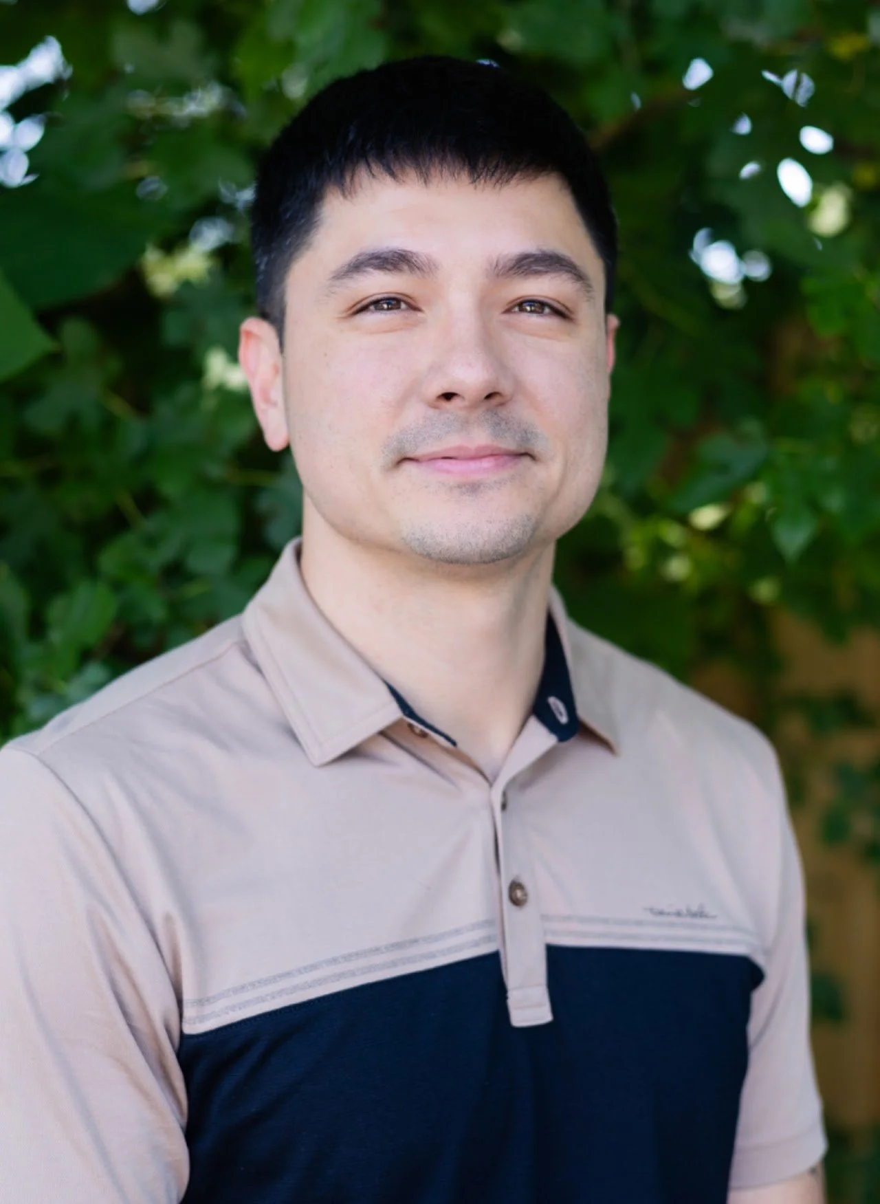 A young man with short black hair, smiling slightly, standing outdoors in front of green foliage, wearing a beige and black collared shirt.