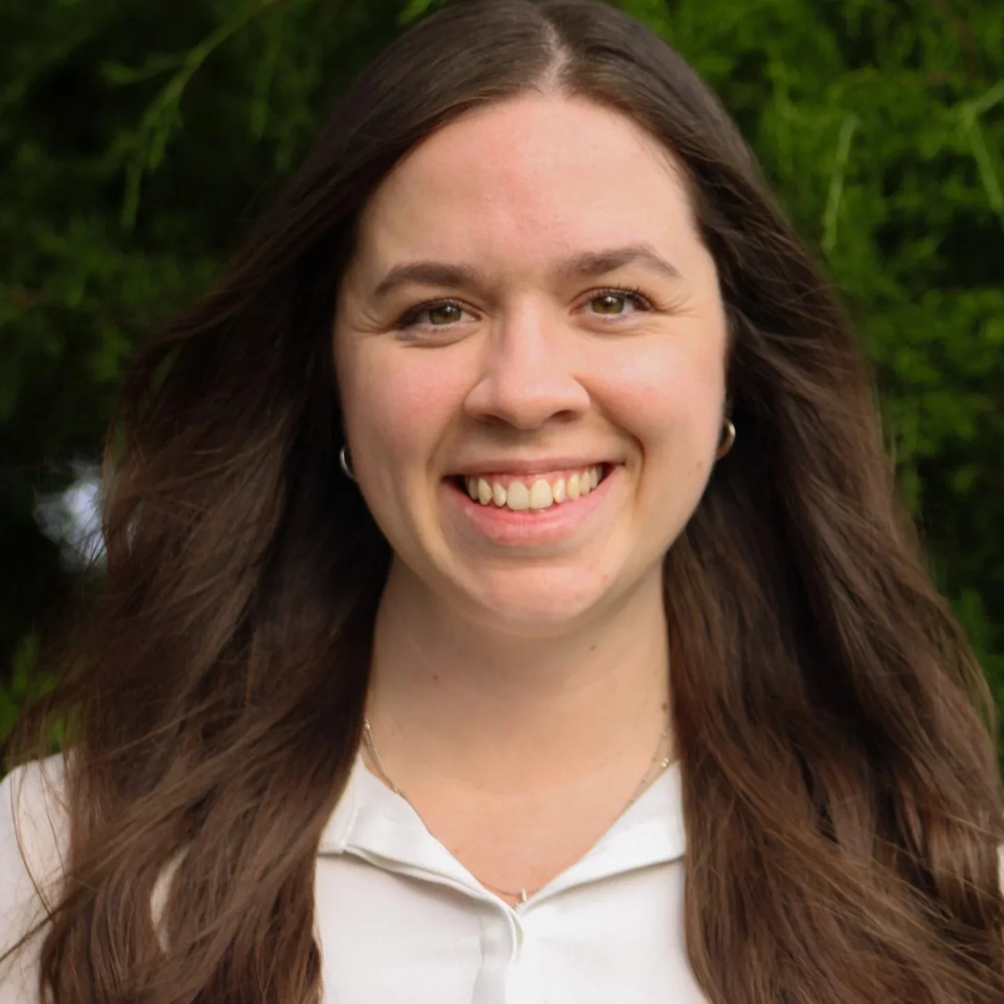 Close-up of a smiling woman with long brown hair, wearing earrings and a white shirt, outdoors with green foliage in the background.