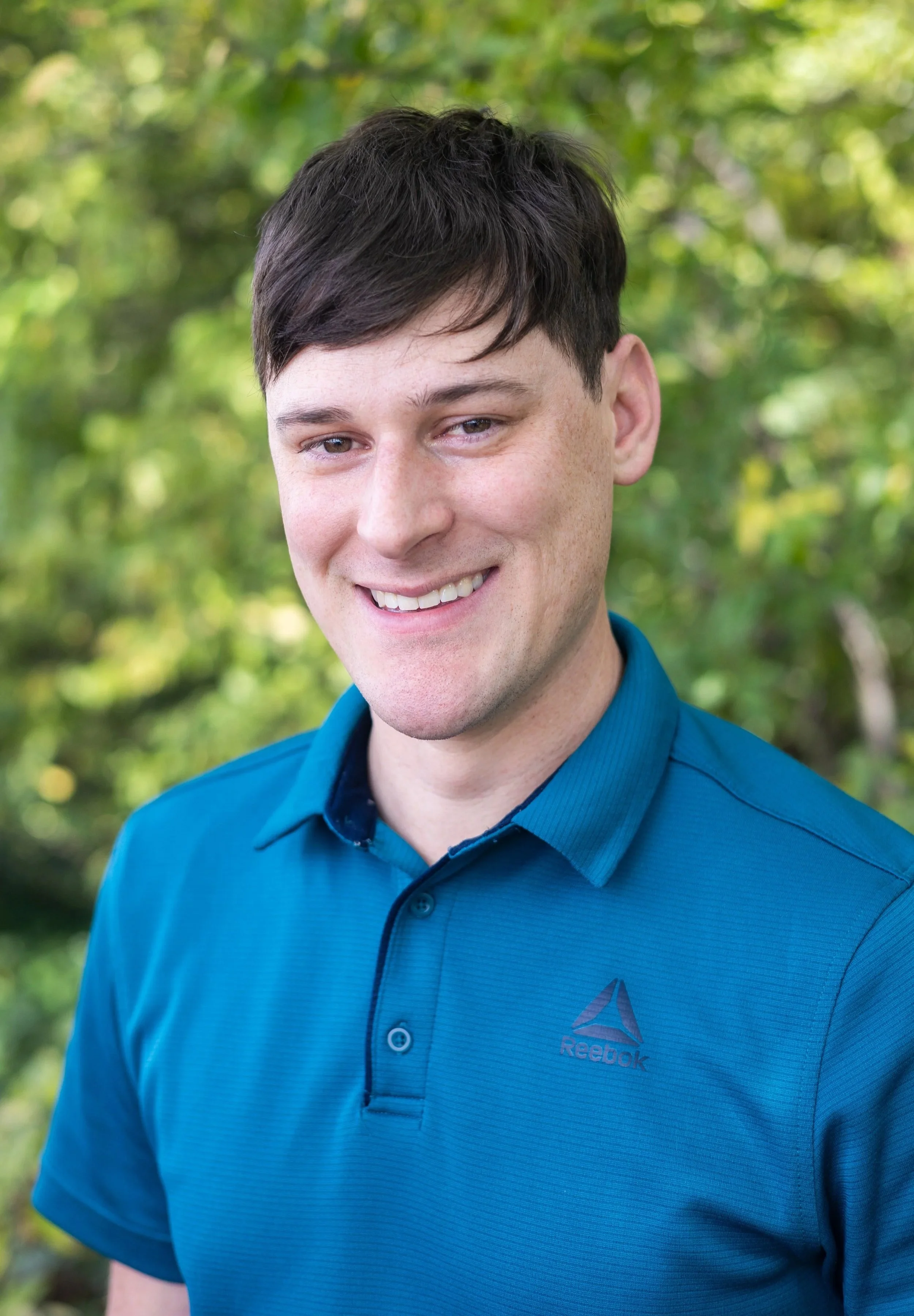 Portrait of a young man with dark hair smiling, wearing a blue Reebok polo shirt, outdoors with green foliage in the background.