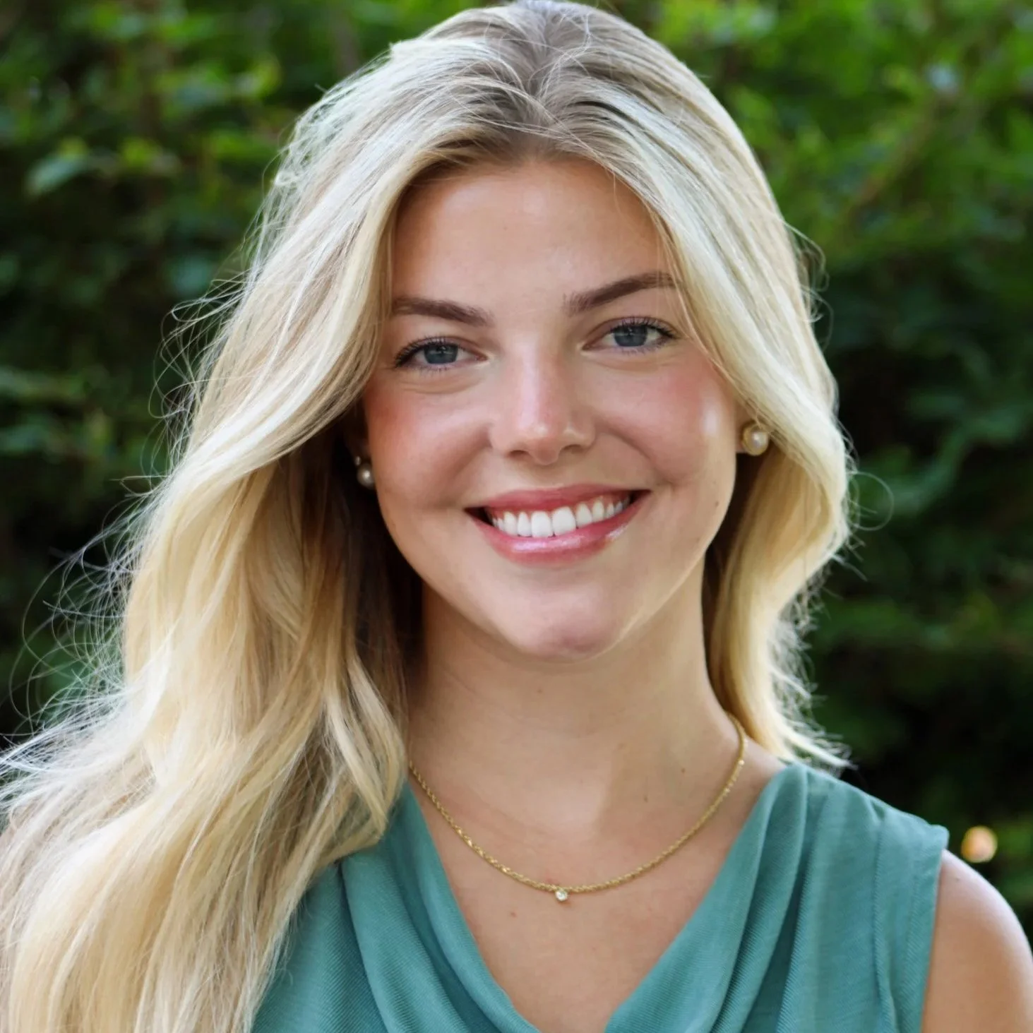 A smiling young woman with long blonde hair, blue eyes, wearing pearl earrings, a gold necklace, and a teal sleeveless top, standing outdoors with green foliage in the background.