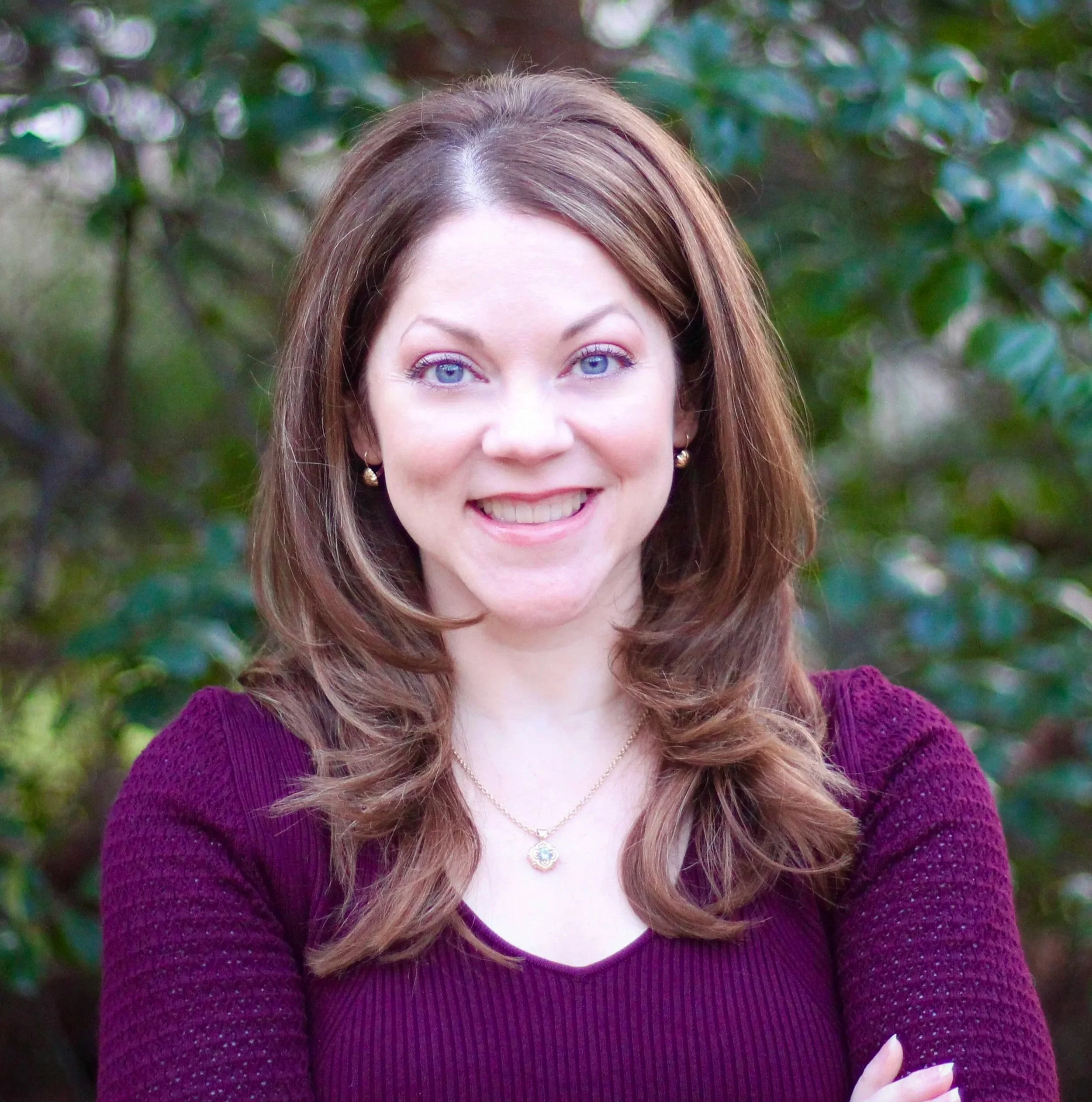 A woman with shoulder-length light brown hair and blue eyes smiling outdoors, wearing a purple top and gold jewelry, with greenery in the background.