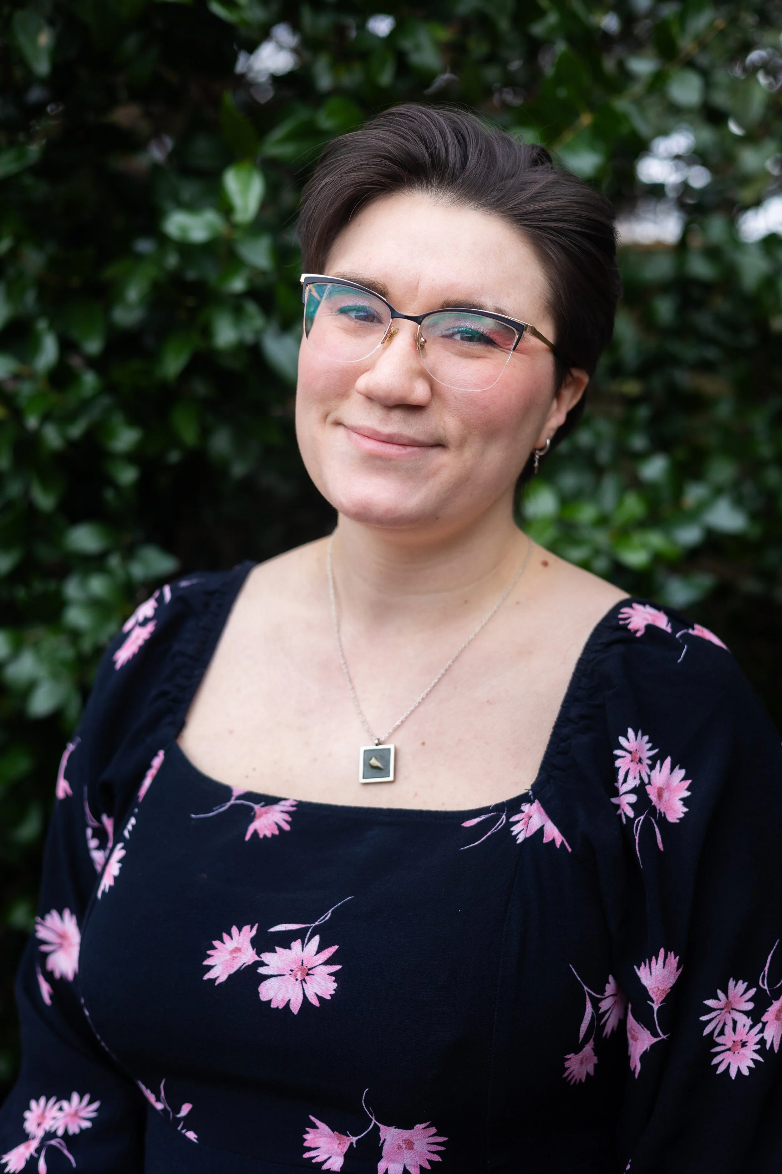 A woman with short dark hair and glasses, wearing a black dress with pink floral patterns and a silver necklace, standing outdoors in front of green foliage.