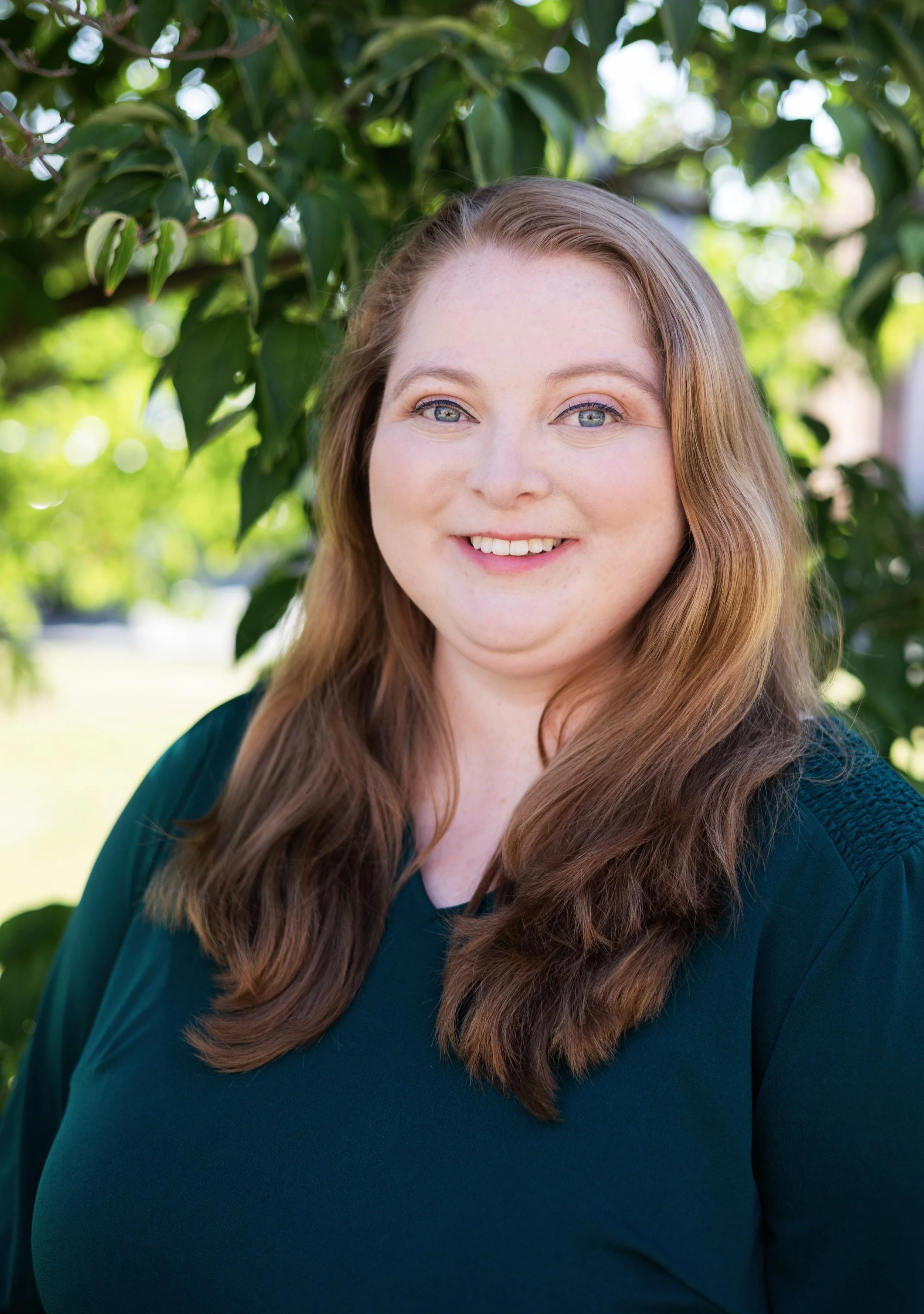 A woman with long brown hair, blue eyes, and a friendly smile standing outdoors among green leaves and trees in daylight.