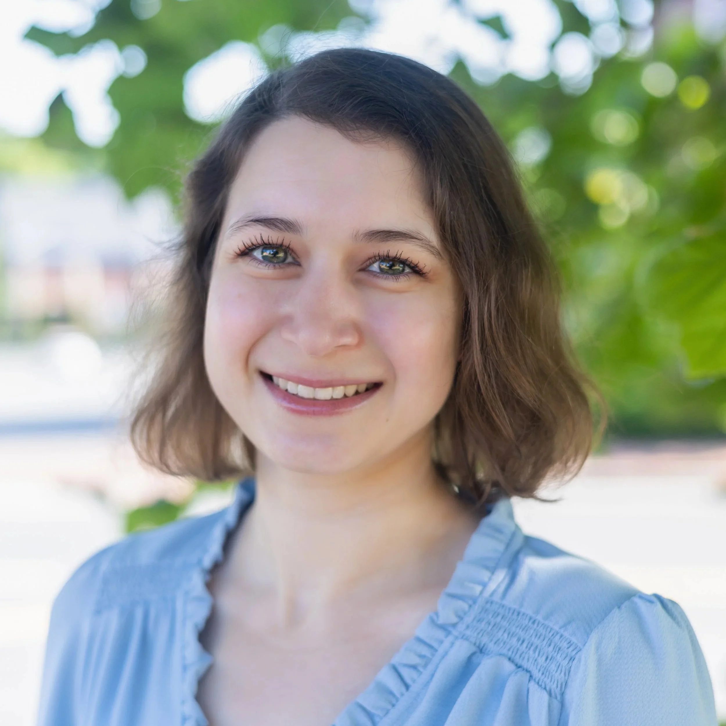 Close-up of a young woman with shoulder-length brown hair, blue eyes, and a bright smile, standing outdoors with green trees and sunlight in the background.