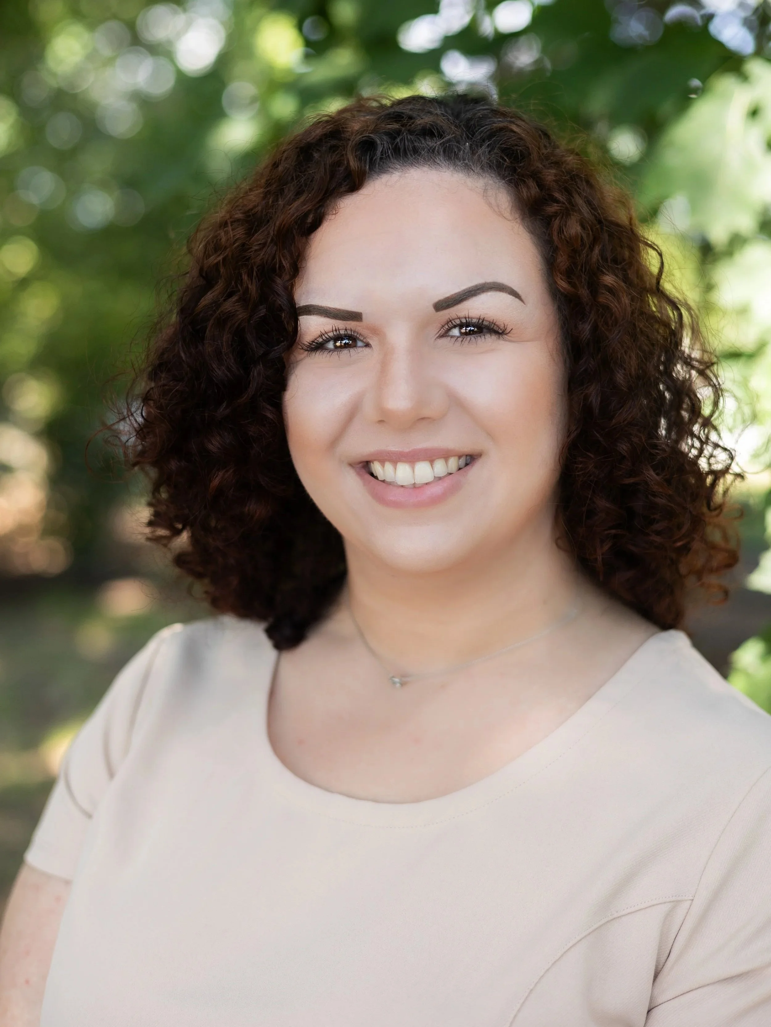Close-up portrait of a woman with curly brown hair, smiling, wearing a light-colored top and a delicate necklace, outdoors with a blurred green leafy background.
