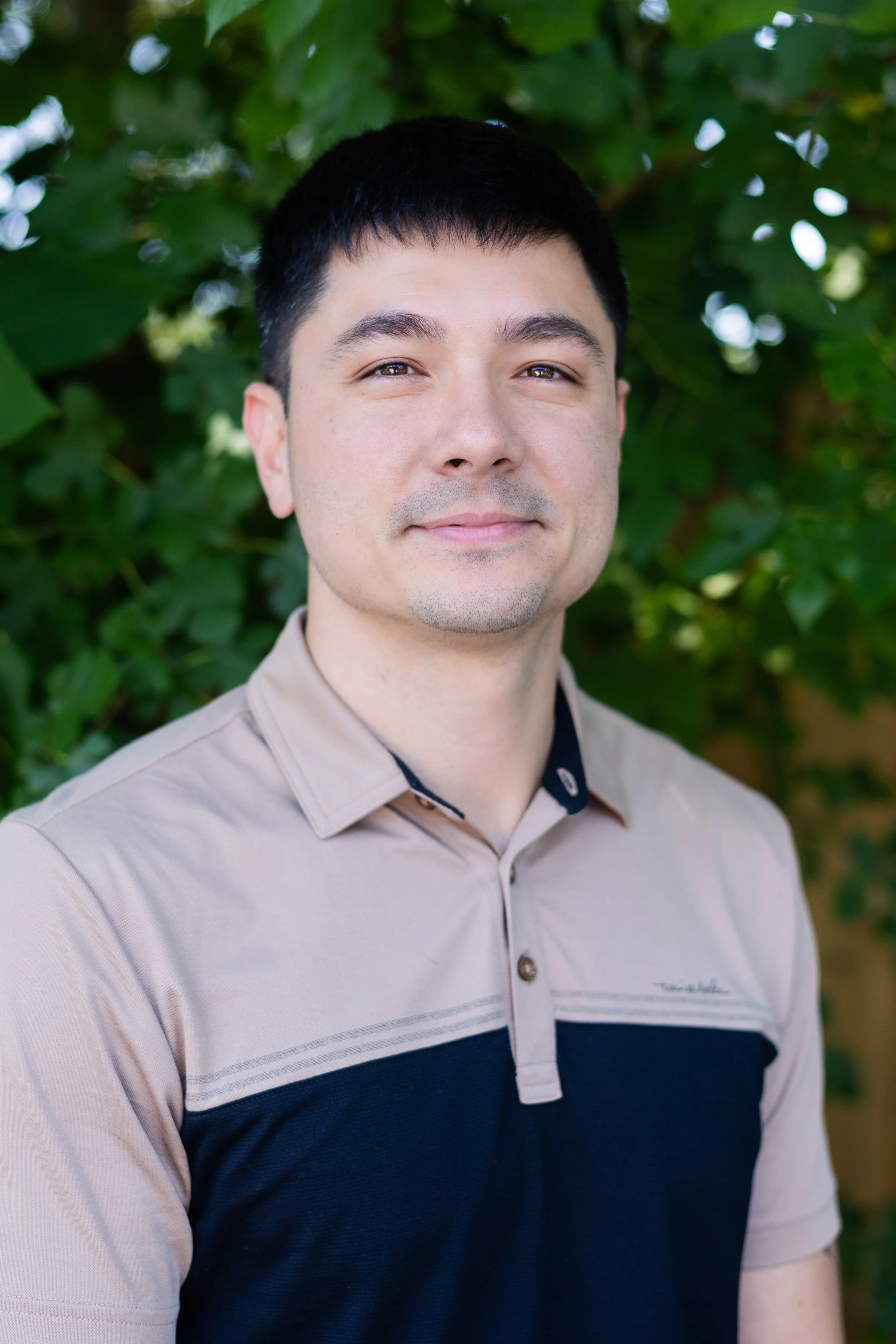 A man with short black hair, wearing a beige and navy polo shirt, standing outdoors in front of green leafy foliage, looking at the camera with a slight smile.