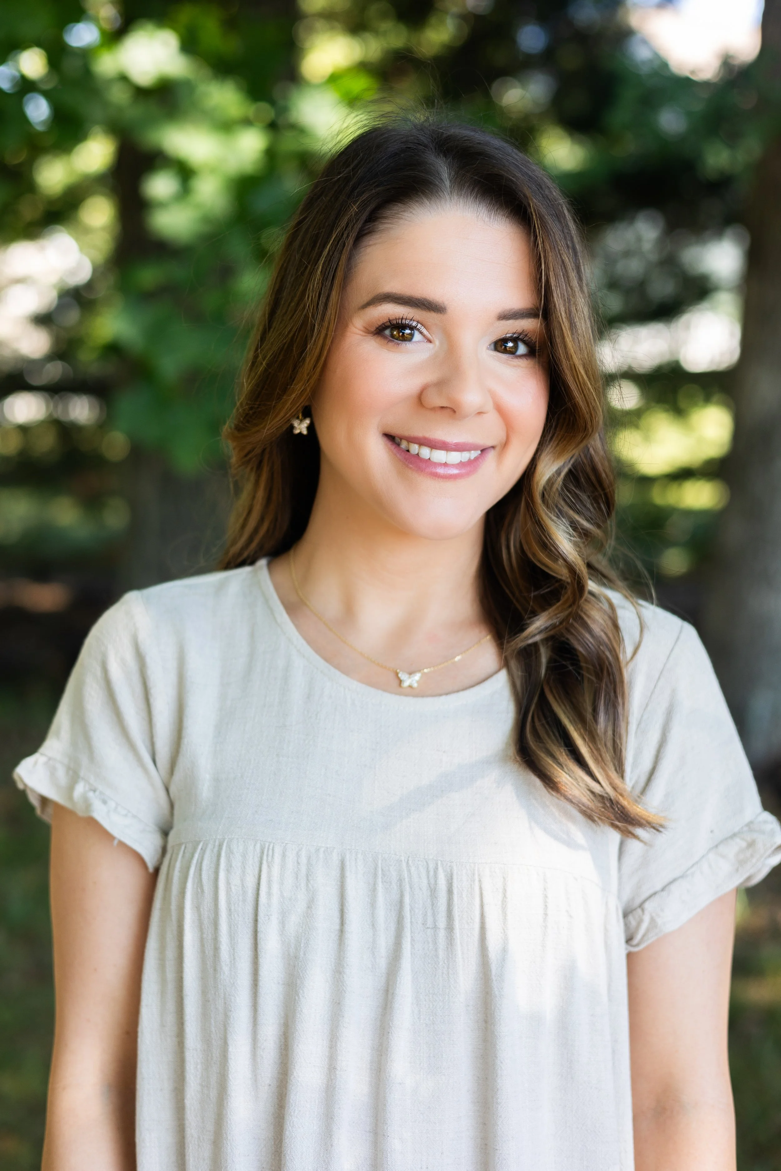 A young woman smiling outdoors with trees in the background, wearing a light-colored dress, butterfly earrings, and a butterfly necklace.