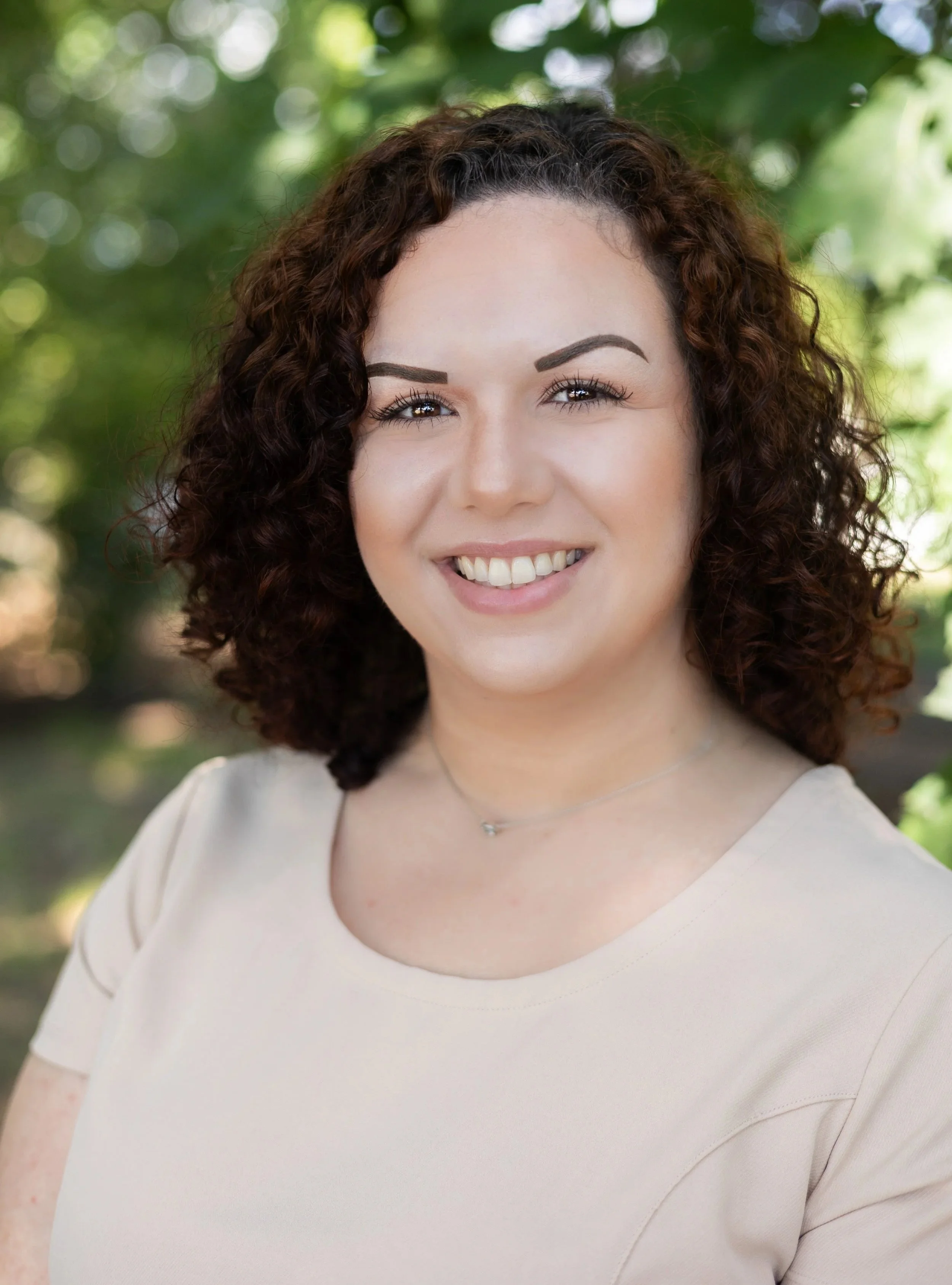 A woman with curly dark hair smiling outdoors with a background of green leaves.