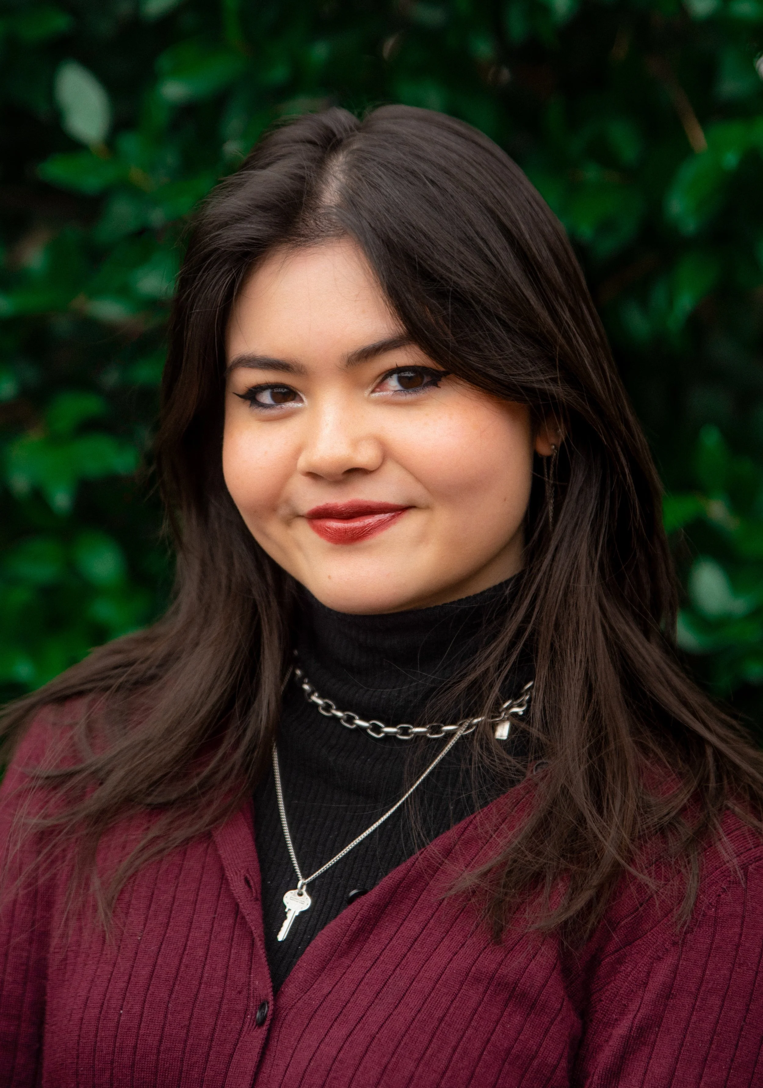 A woman with long dark hair and makeup, wearing a black turtleneck, layered silver necklaces, and a burgundy button-up top, smiling outdoors in front of green foliage.