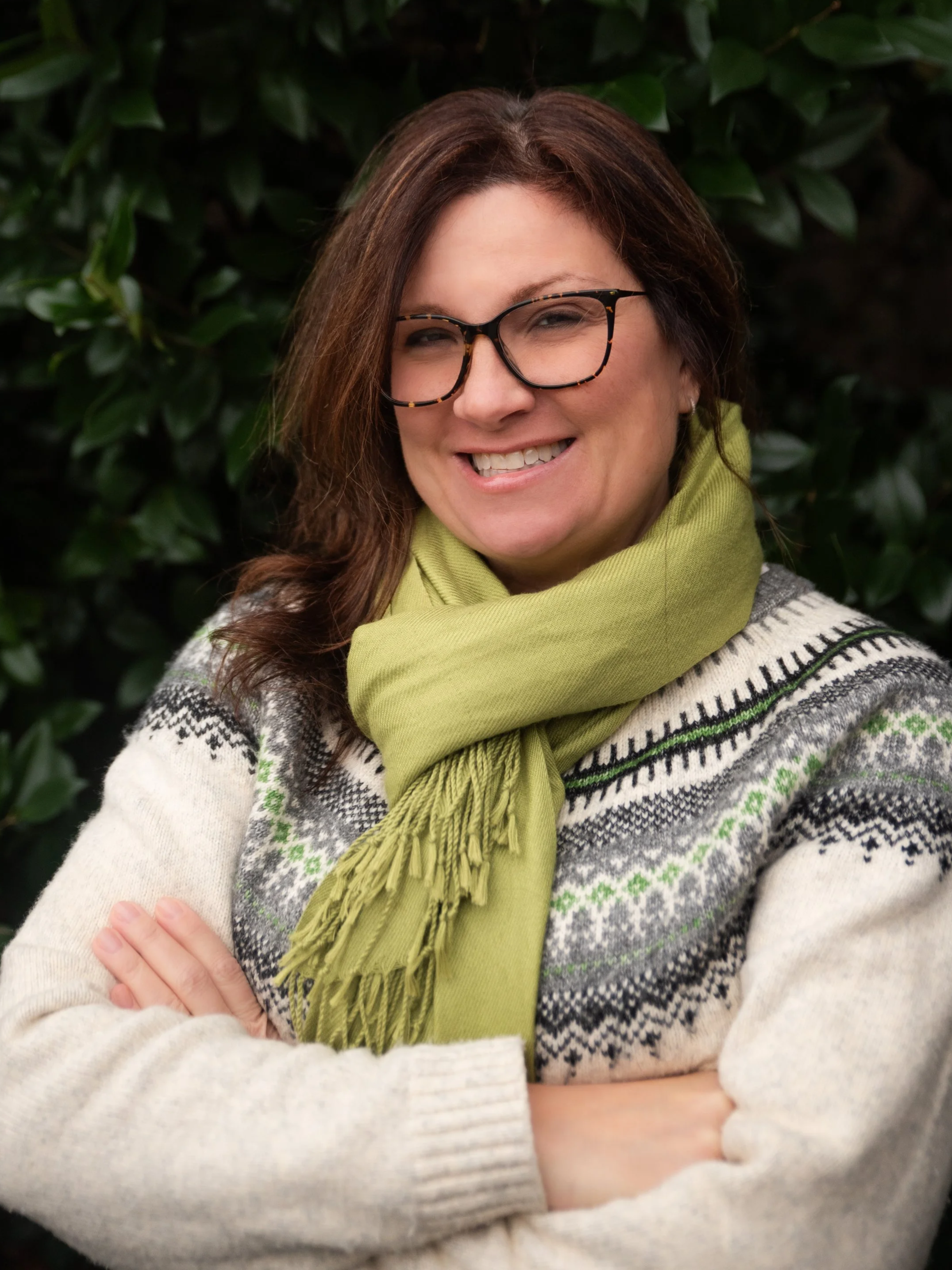 A smiling woman with glasses, wearing a patterned sweater and a green scarf, crossing her arms in front of green foliage.