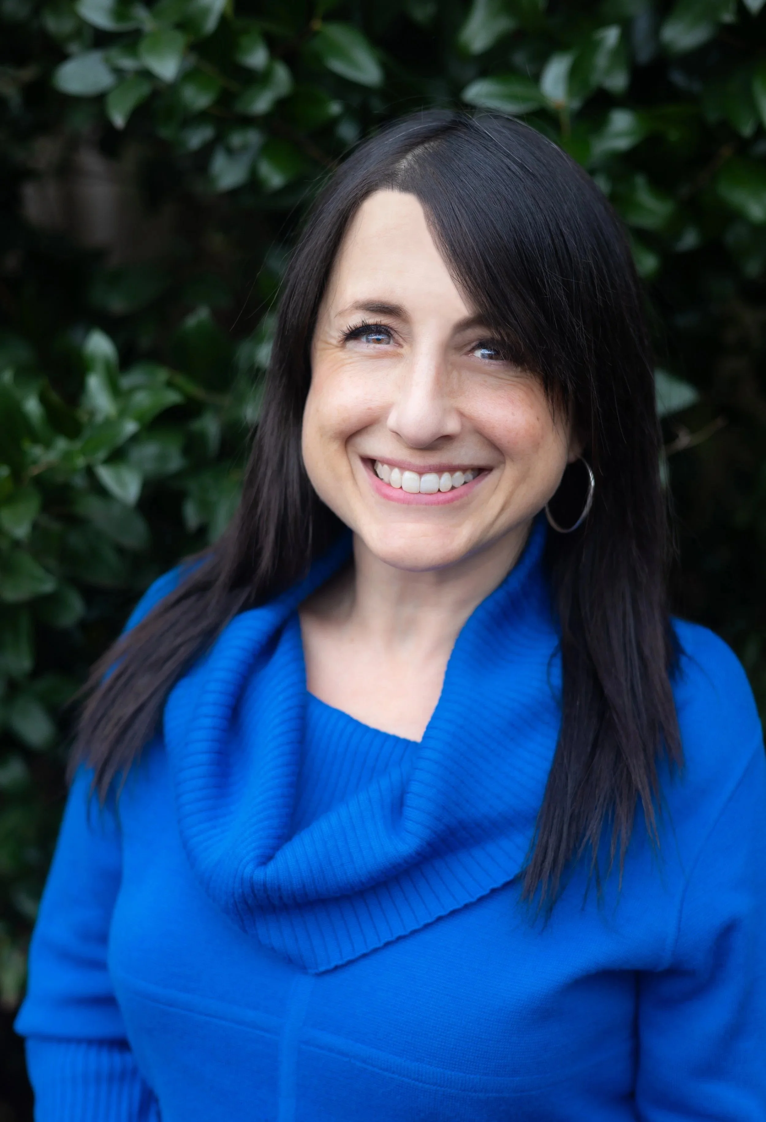A woman with dark hair, blue eyes, and hoop earrings smiling in front of green foliage, wearing a royal blue cowl-neck sweater.