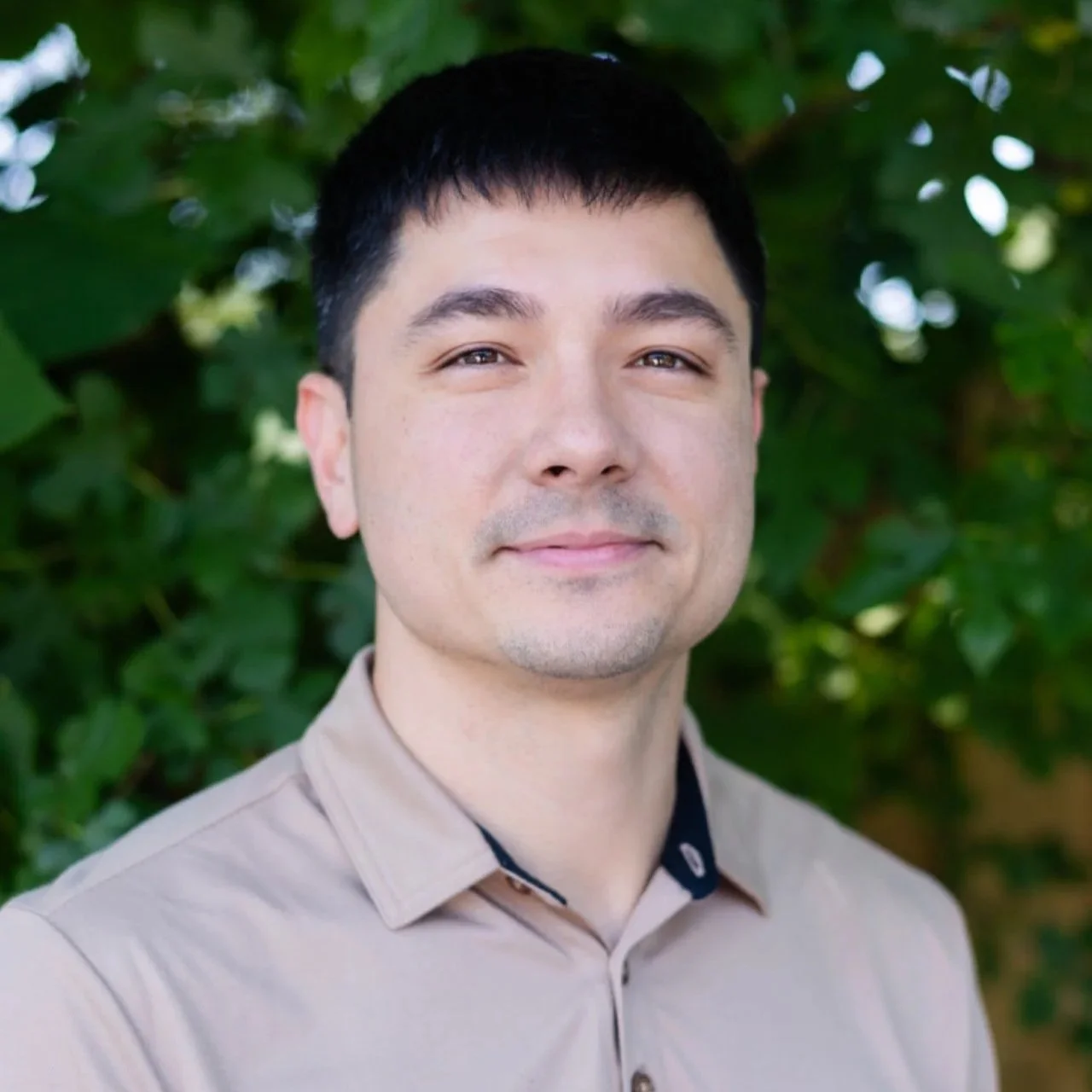 Close-up portrait of a man with short black hair, wearing a beige collared shirt, standing outdoors with green foliage background.