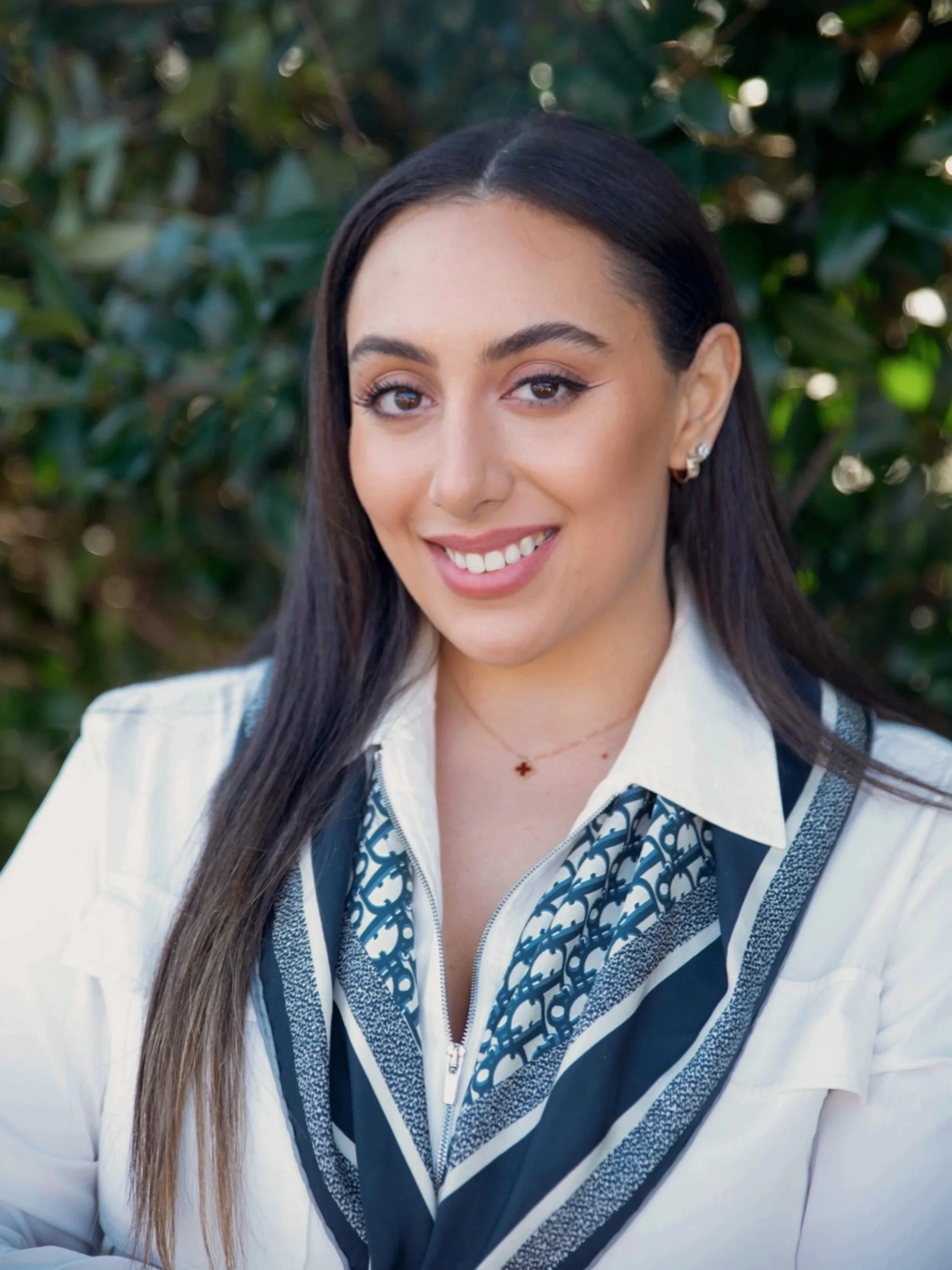 A woman with long dark hair smiling outdoors, wearing a white top with a patterned scarf, and jewelry including earrings and a delicate necklace.