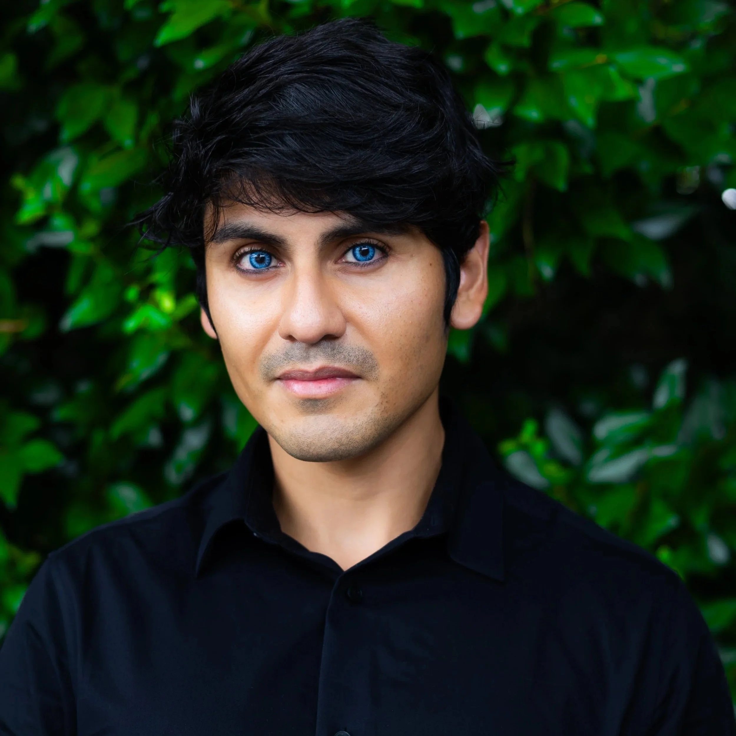 A man with black hair and bright blue eyes, wearing a black shirt, standing against a background of green leaves.