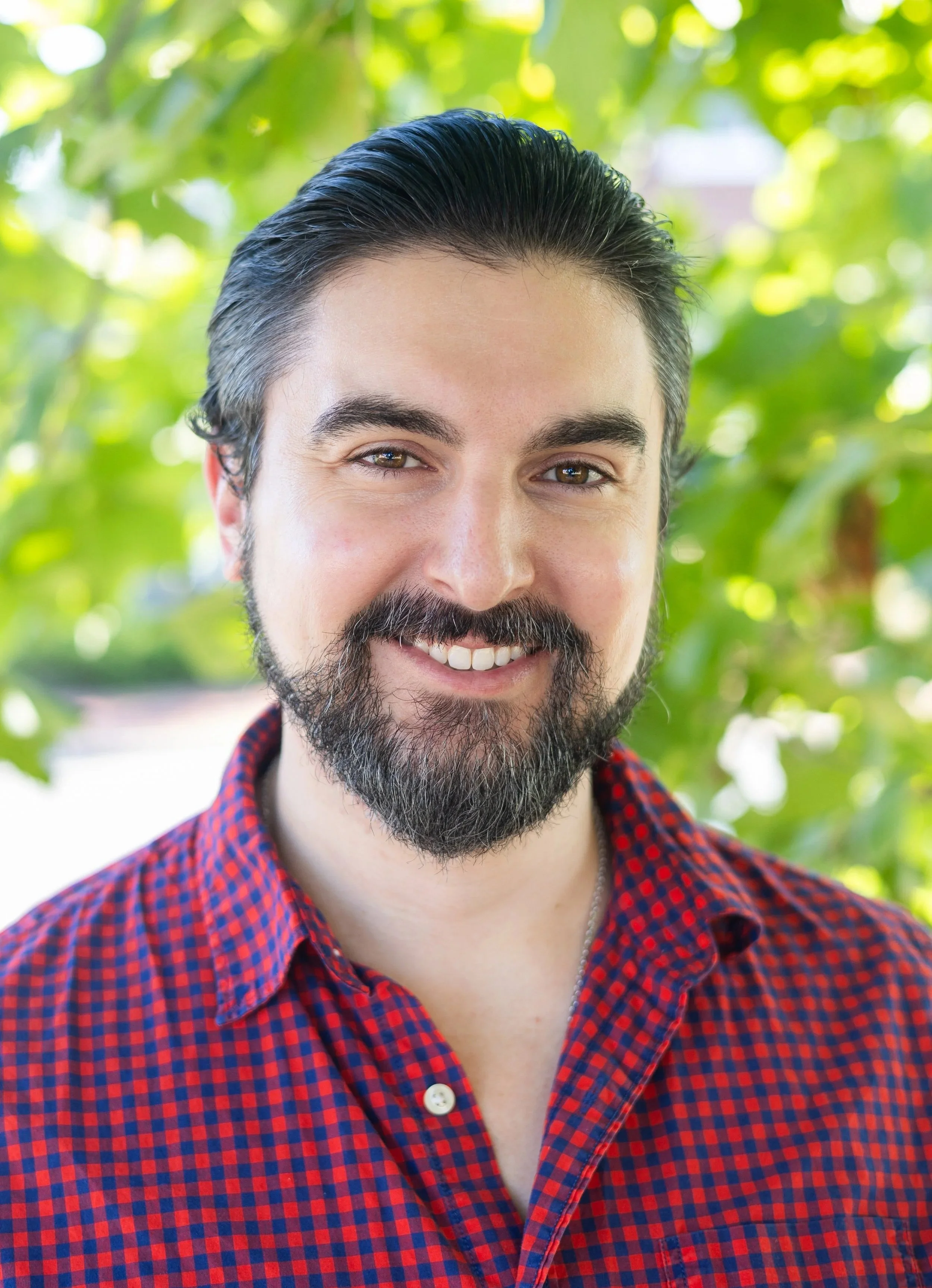 A smiling man with dark hair, a beard, and mustache, wearing a red and blue checkered shirt, standing outdoors with green foliage in the background.