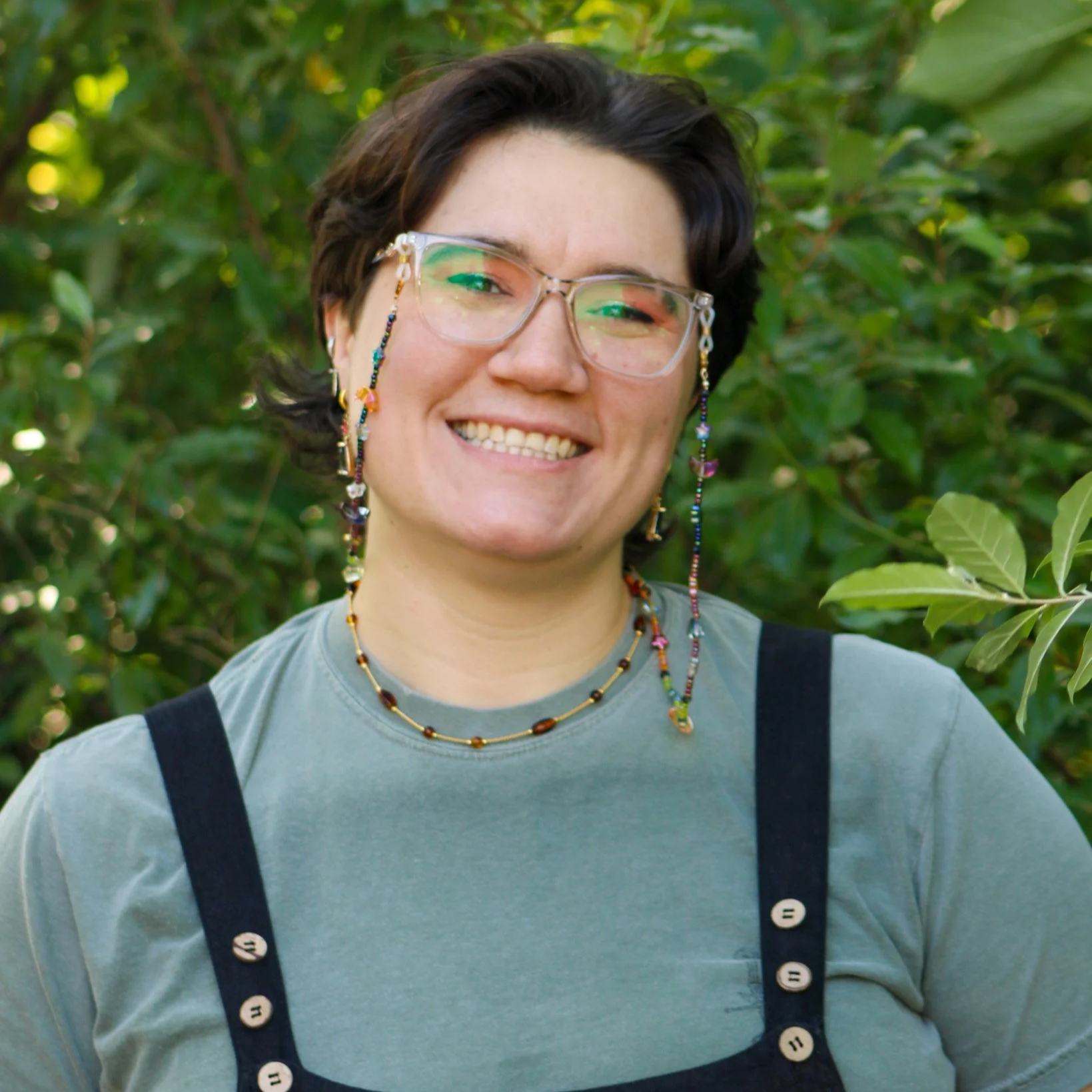 A smiling person with short dark hair wearing glasses with beaded chains, layered necklaces, a gray T-shirt, and black suspenders, standing outdoors in front of green foliage.