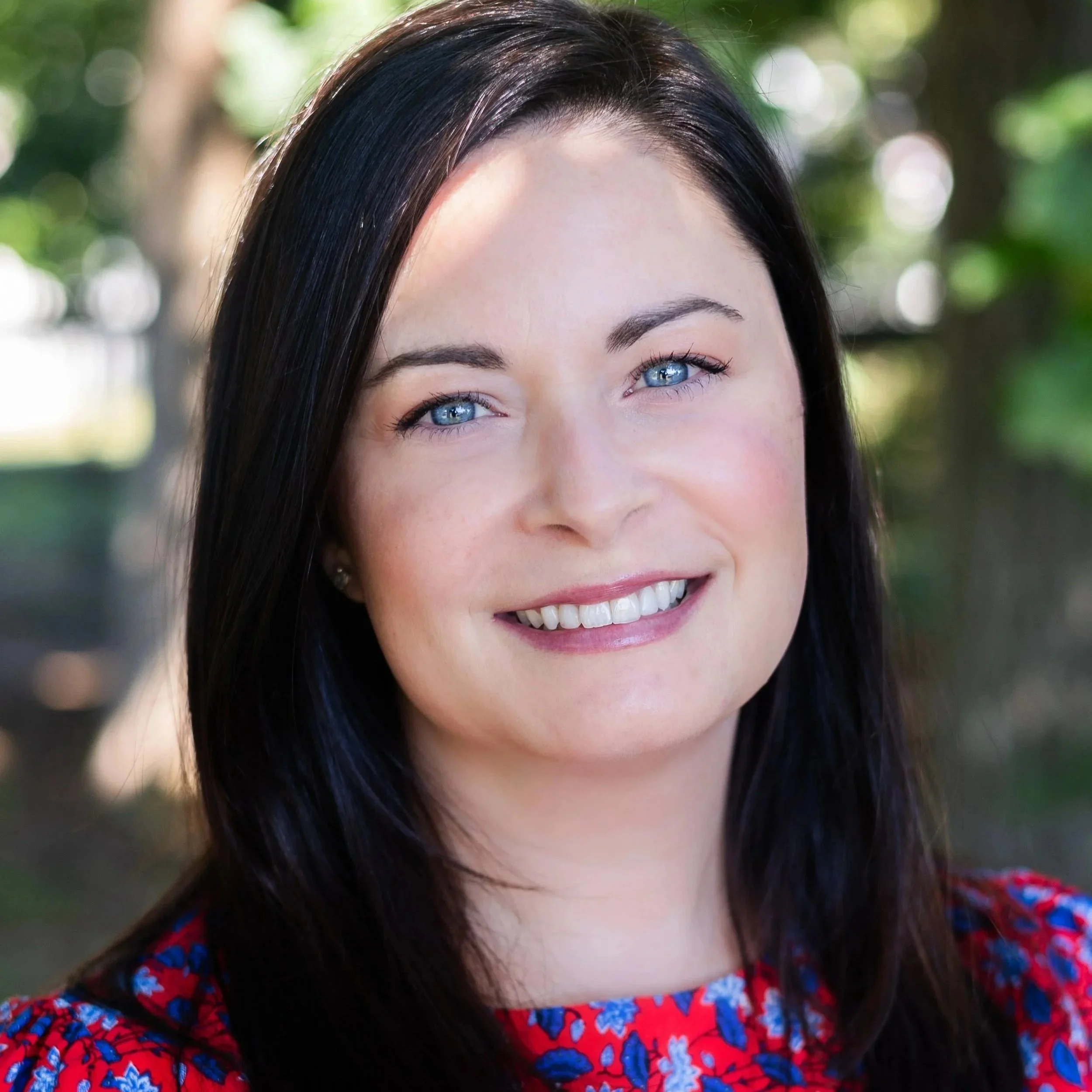 Close-up of a woman with blue eyes and dark brown hair smiling outdoors with a blurred green background.