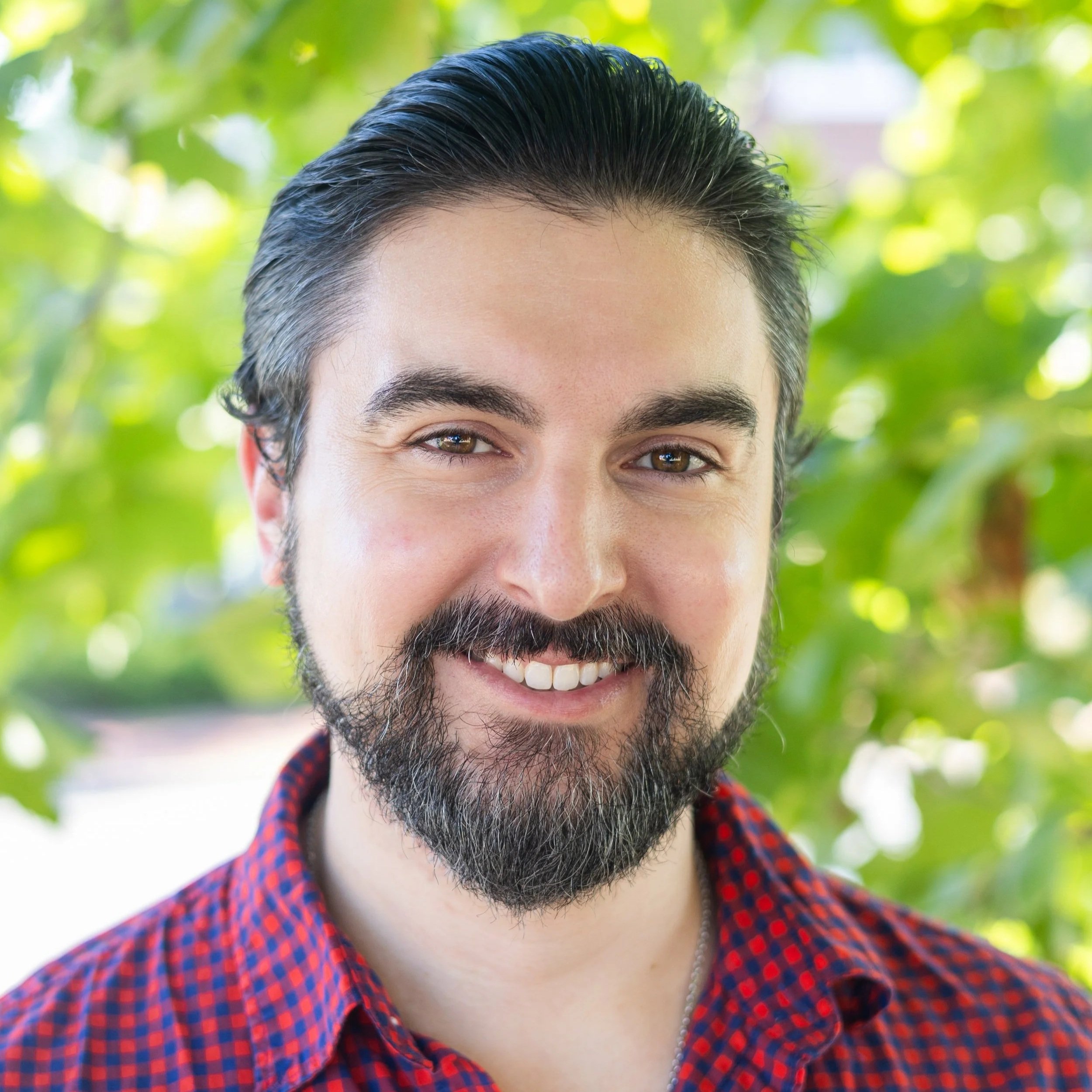 Close-up of a smiling man with dark hair and a beard, wearing a red and blue checkered shirt, outdoors with green foliage in the background.