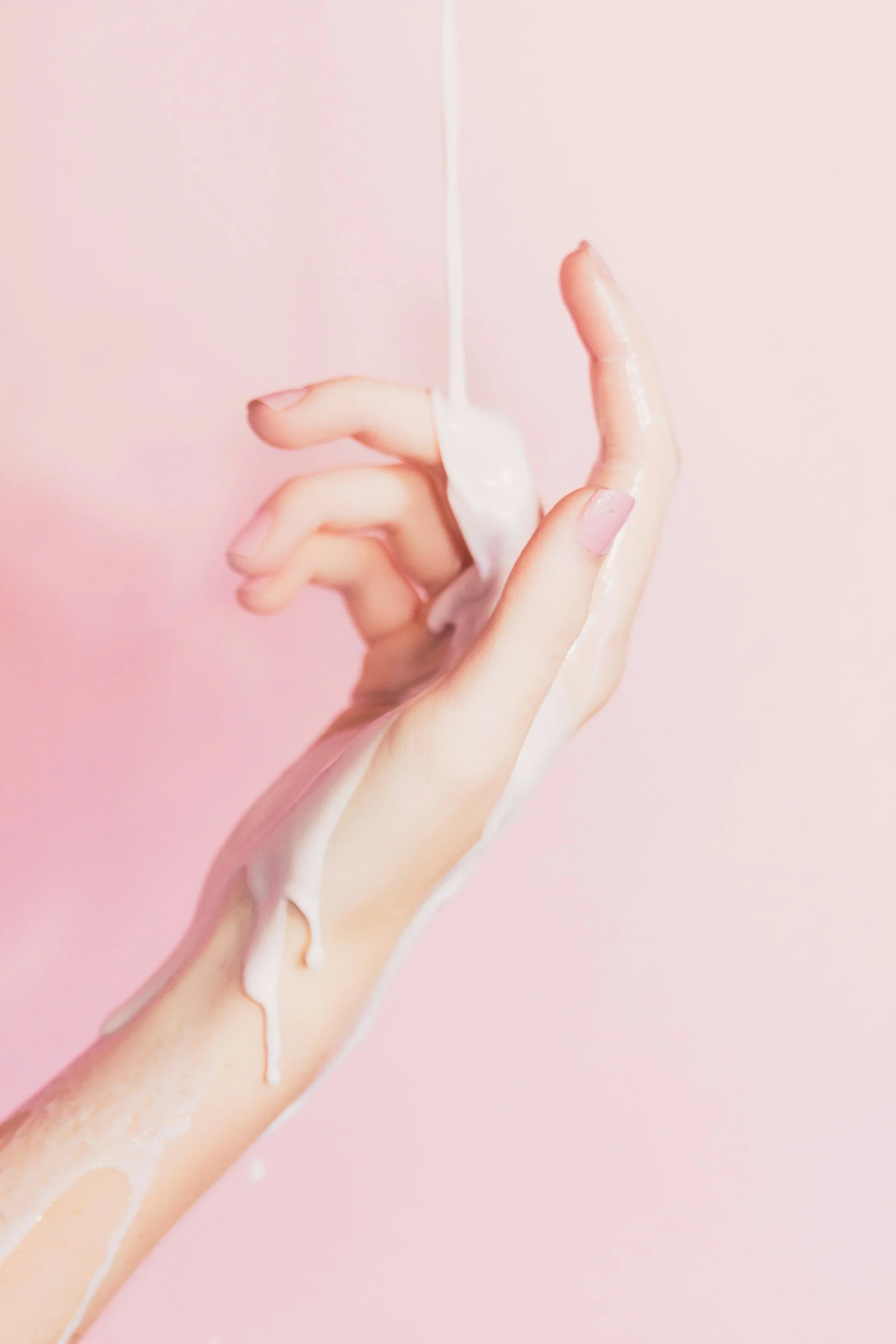 A hand with light pink nail polish is shown with a white liquid, possibly lotion or cream, being poured over it against a pink background.