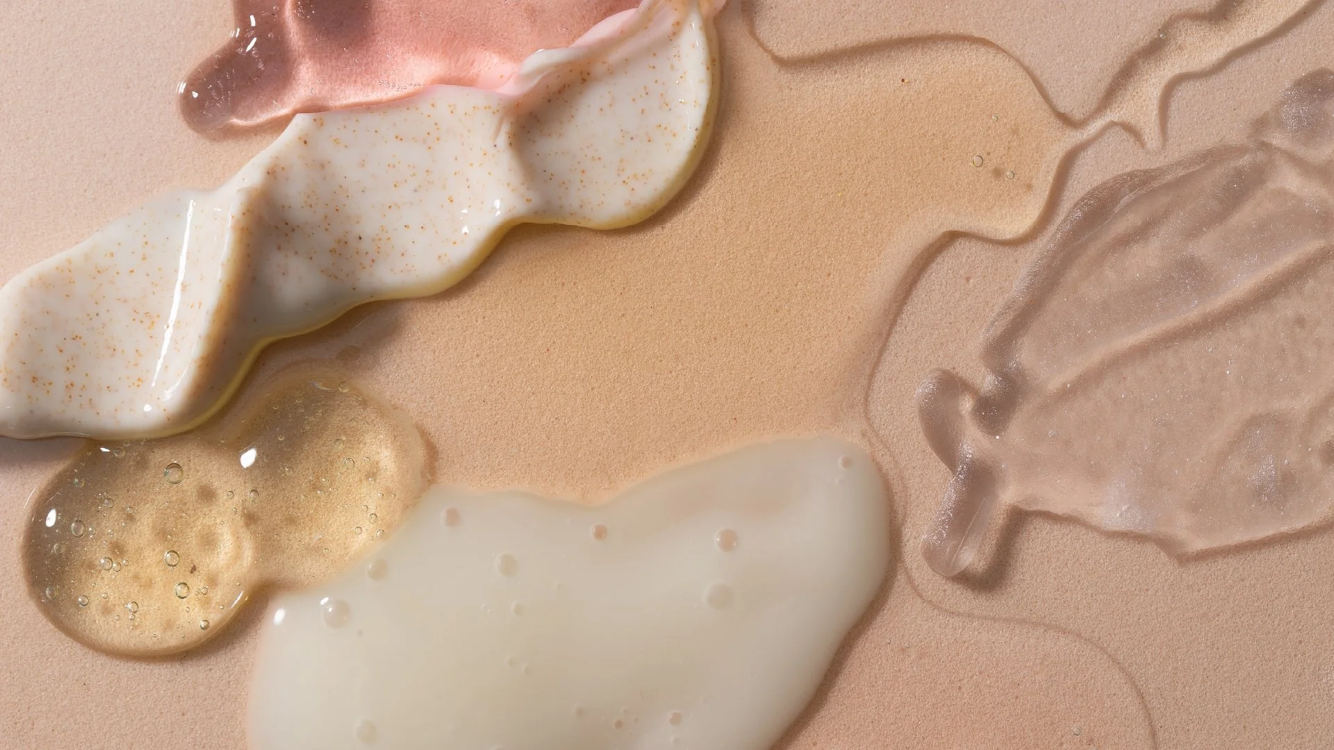 Various liquids with textures and colors on a peach-colored surface, including white, beige, pink, and translucent substances.
