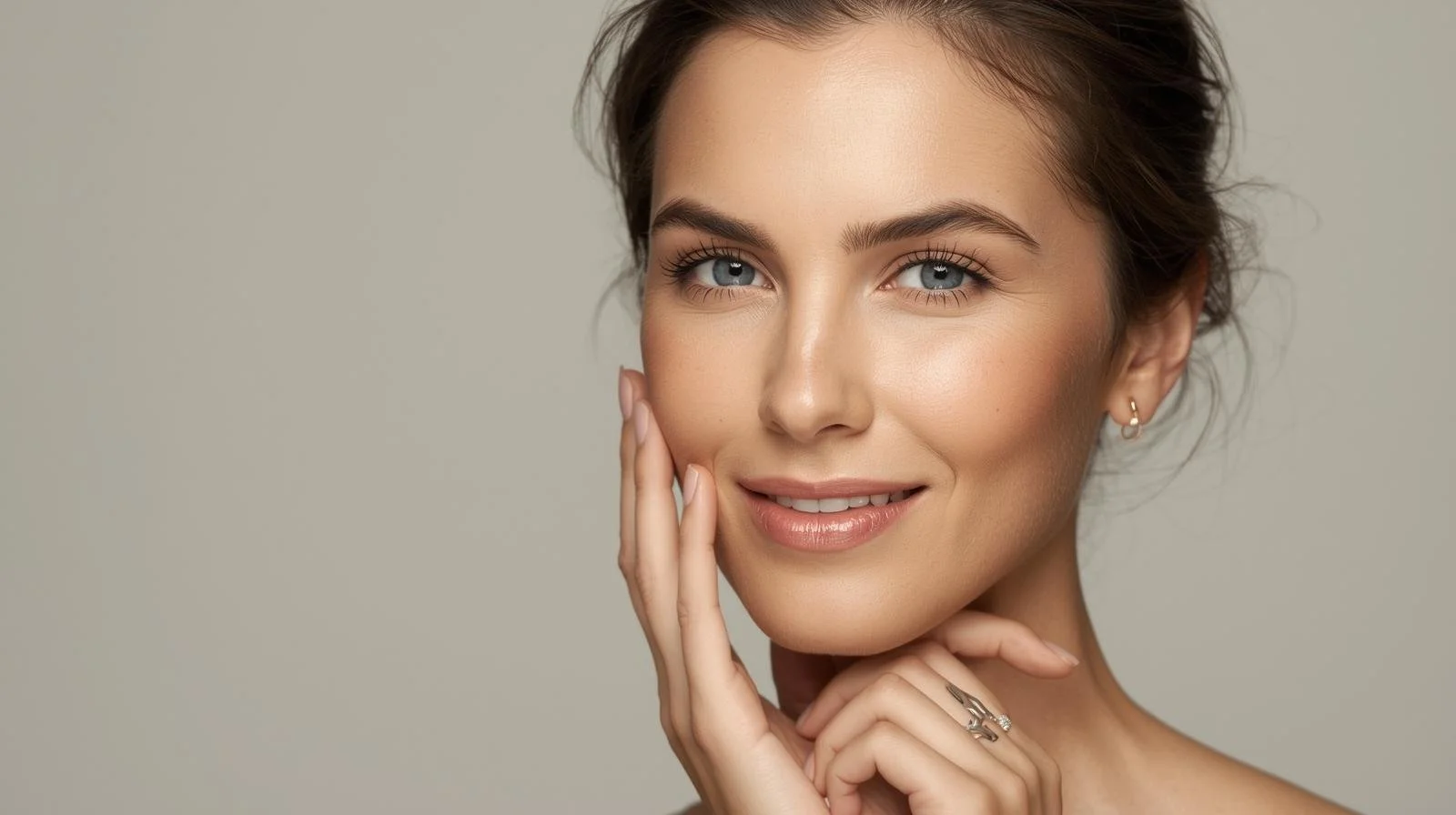 Close-up of a smiling woman with blue eyes and brunette hair, gently touching her face with her right hand.