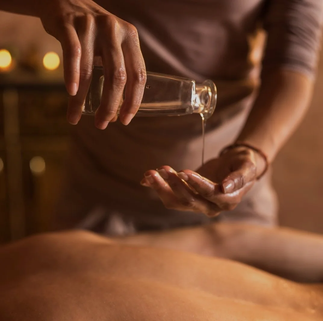 Person pouring massage oil from a glass bottle onto a person's back during a massage in a dimly lit room.