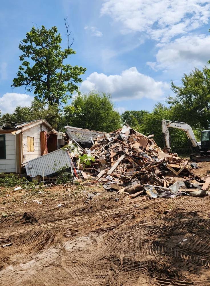 A destroyed house with scattered debris, an excavator on the right, and a partly cloudy sky with green trees in the background.