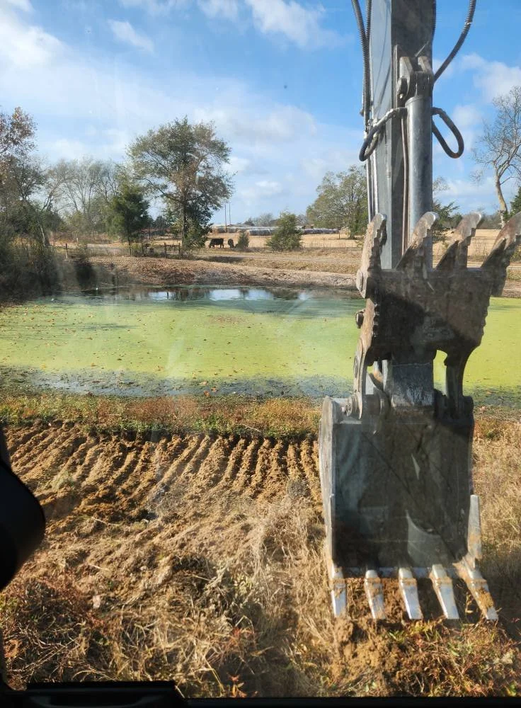 View through a construction vehicle window showing an excavator bucket in the foreground, a small pond with algae, trees, and a farm landscape with horses in the background.