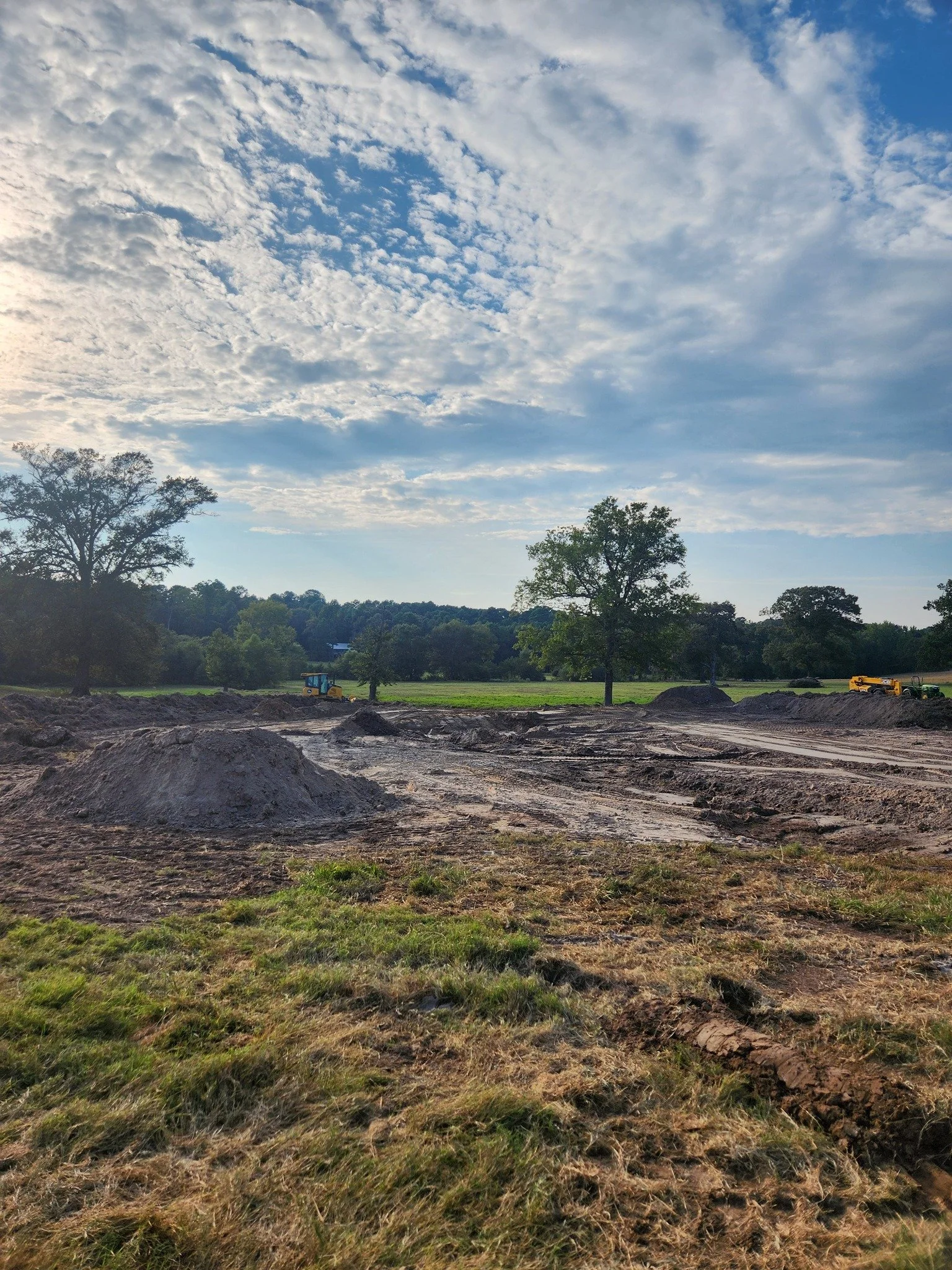 Construction site with piles of dirt, dirt tracks, and trees in the background under a partly cloudy sky.