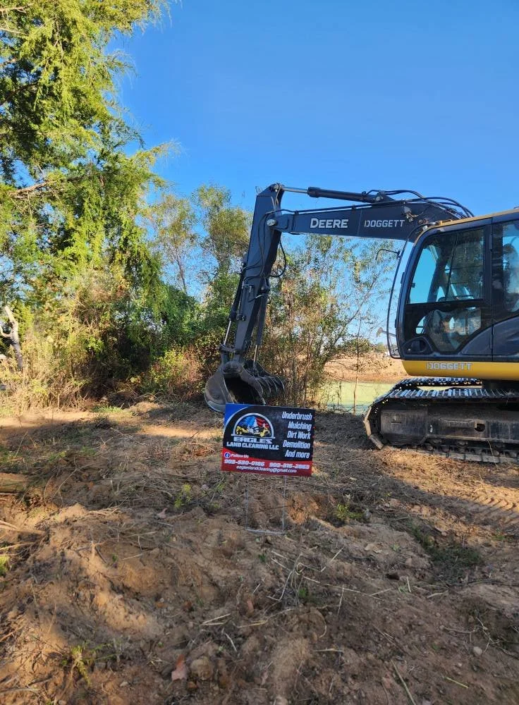 A yellow and black John Deere excavator operating on dirt near trees, with a sign that reads "Underground Mulching, Dirt Work, Demolition, And More" in front of it.